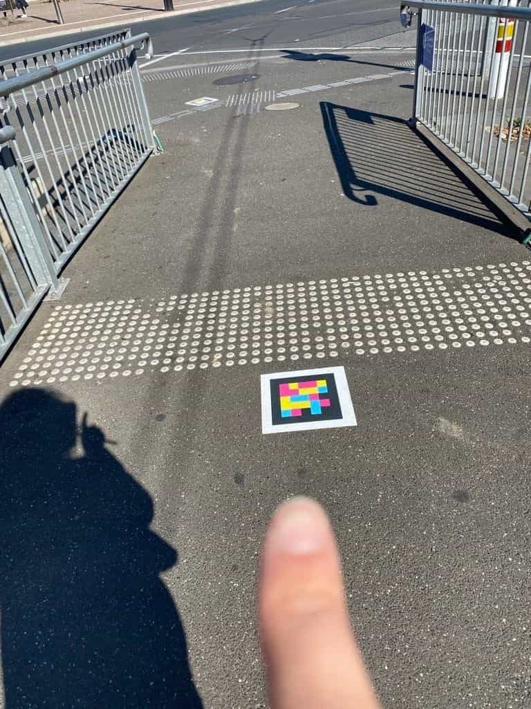Código NaviLens enmarcado en blanco pegado en el suelo de asfalto justo antes de una franja podotáctil de botones blancos en relieve, en la entrada peatonal a un andén con barandillas metálicas a ambos lados