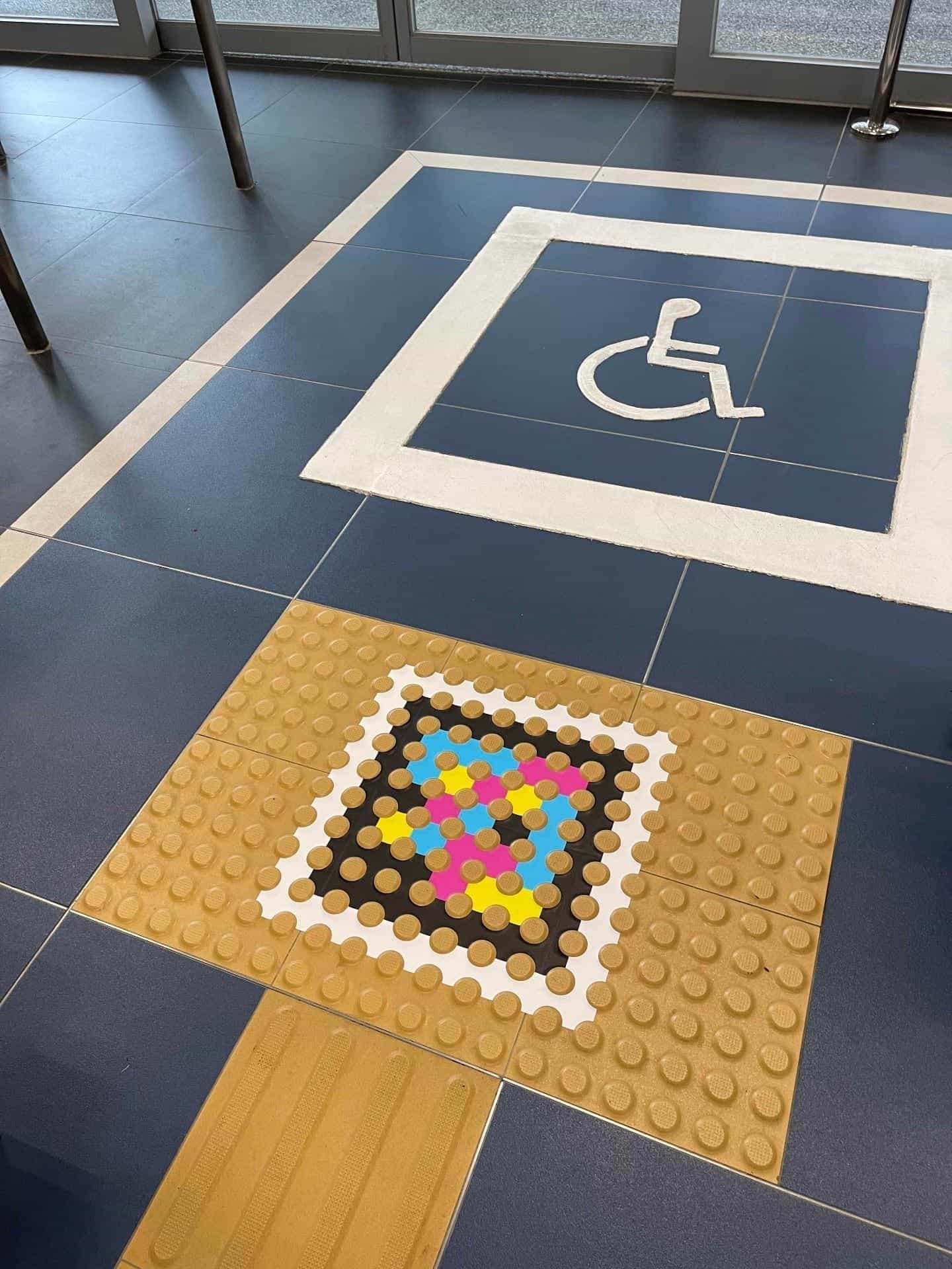 Dark blue floor with a white wheelchair pictogram and a mustard tactile panel with an inset NaviLens code