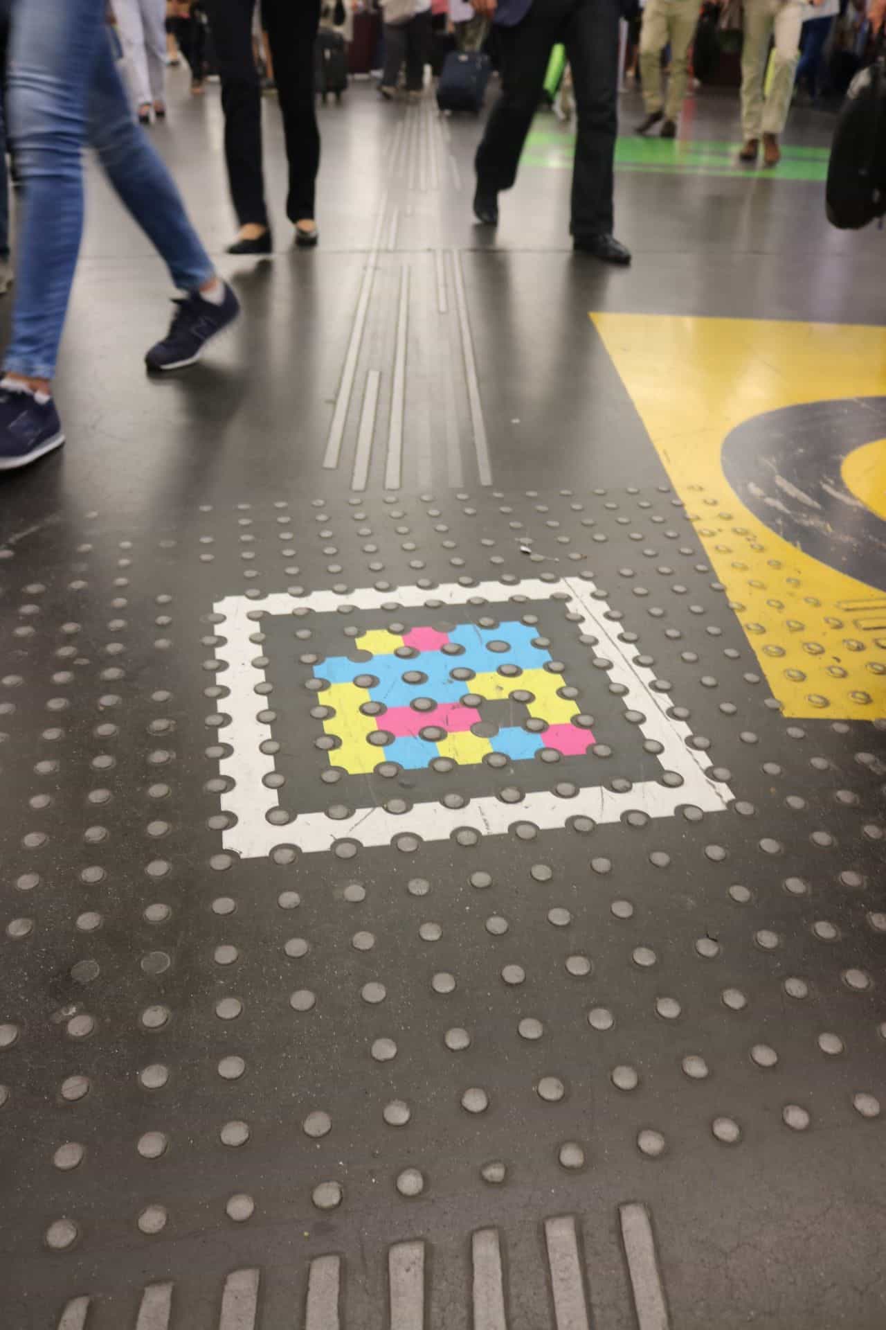 Overhead view of the Atocha concourse floor at rush hour: a white-framed NaviLens code is embedded in a tactile paving strip of studs, next to yellow signage; travelers with suitcases walk in the background.