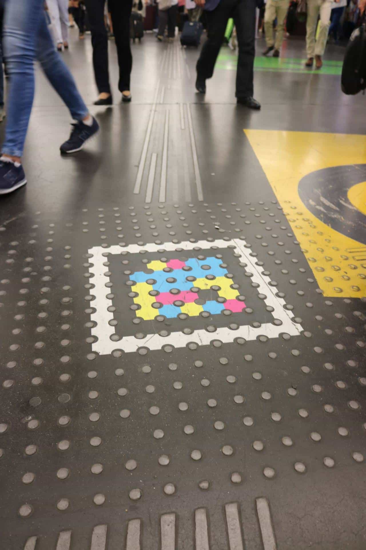 Overhead view of the Atocha concourse floor at rush hour: a white-framed NaviLens code is embedded in a tactile paving strip of studs, next to yellow signage; travellers with suitcases walk in the background.