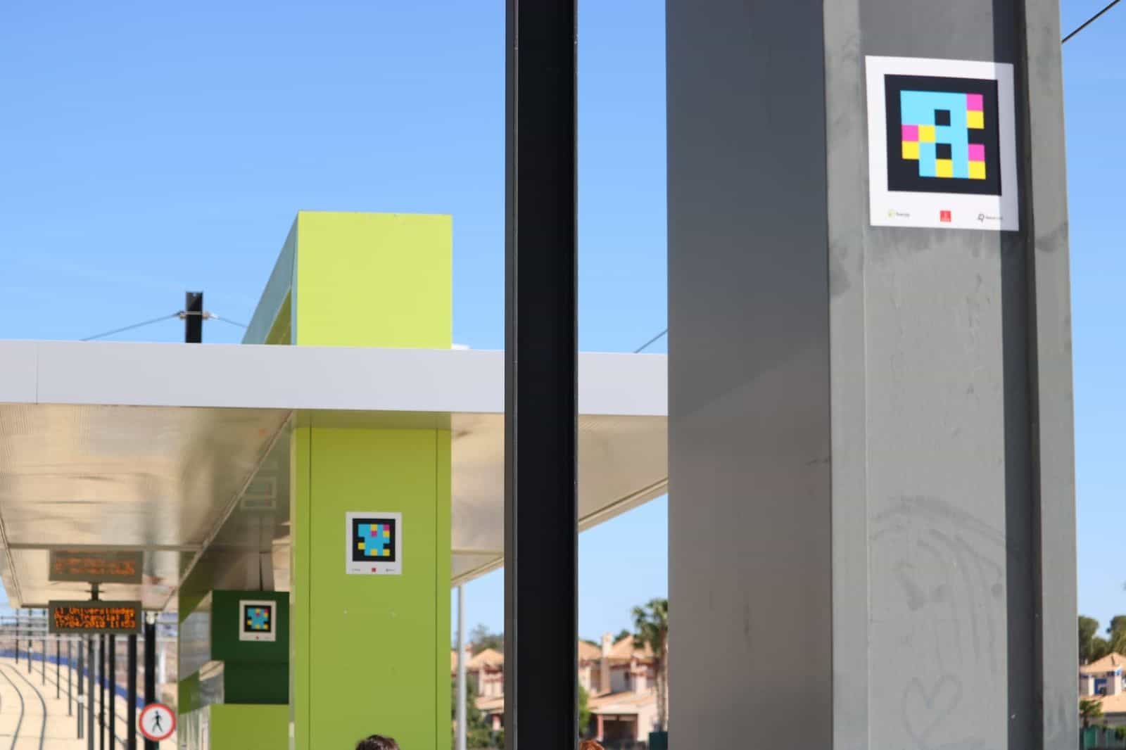 Persona che inquadra un codice NaviLens collocato in cima a una pensilina del Tranvía de Murcia, con lo schermo LED dei prossimi arrivi in fondo