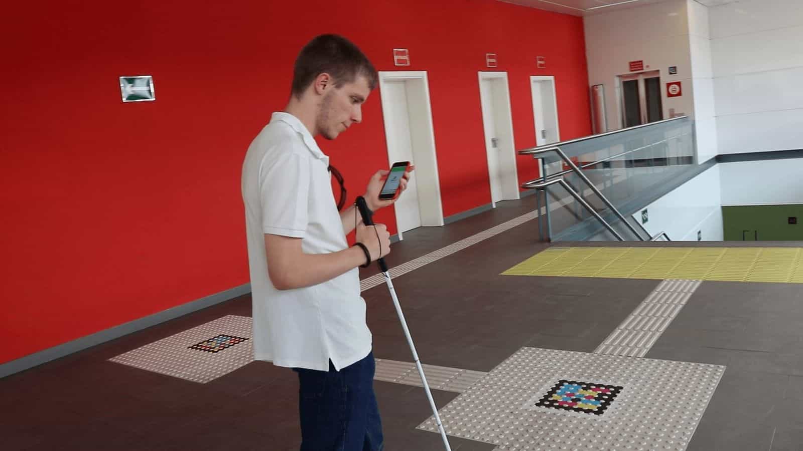 Station corridor with a red wall and dark floor: two NaviLens codes embedded in tactile paving strips mark the route to the stairs; a young user with a white cane checks the NaviLens GO app on their phone.