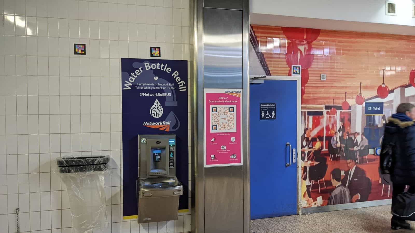 Two small NaviLens codes on the white-tiled wall next to the Network Rail «Water Bottle Refill» fountain and the blue accessible toilet door at Euston