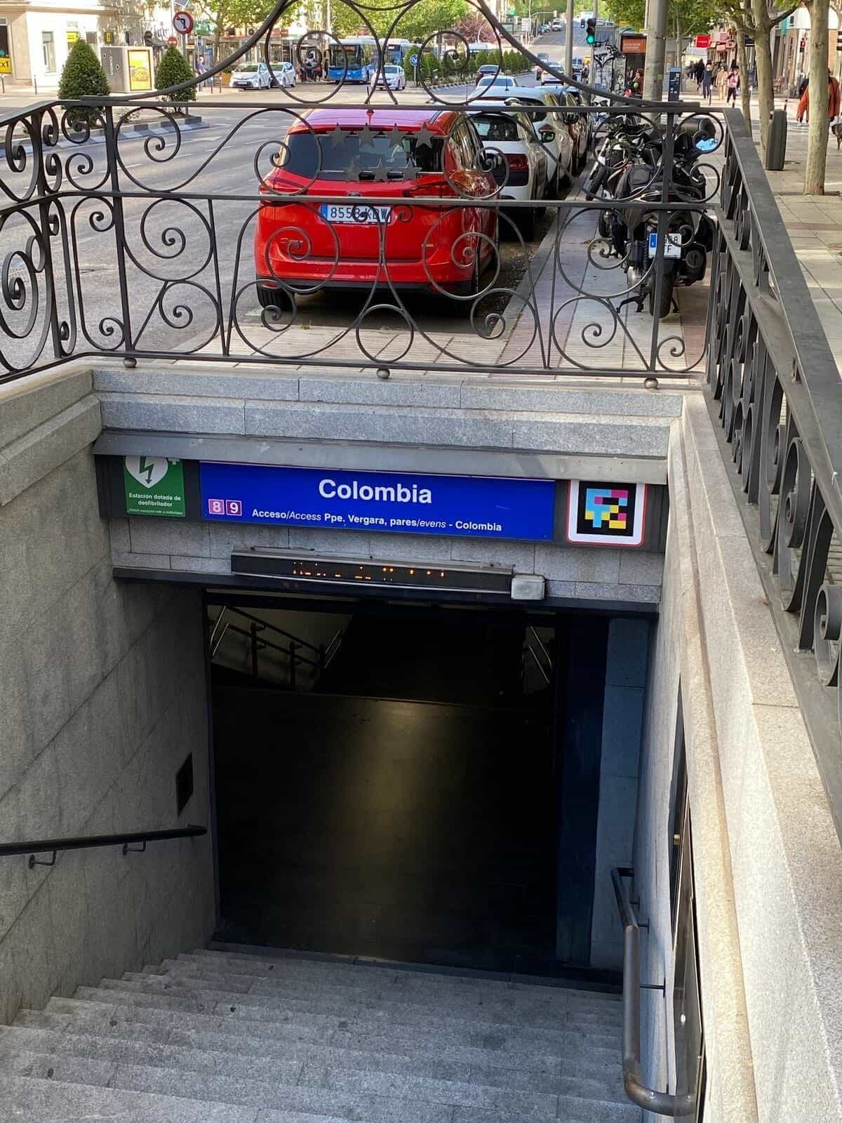 Detail des Zugangs zu Colombia von der Straße: unter dem schmiedeeisernen Geländer das blaue Identifikationsschild des Bahnhofs „Colombia“, die Symbole der Linien 8 und 9, der Hinweis „Acceso/Access Ppe. Vergara, gerade Zahlen — Colombia“ und rechts ein quadratischer farbiger NaviLens-Code auf seiner weißen Raute