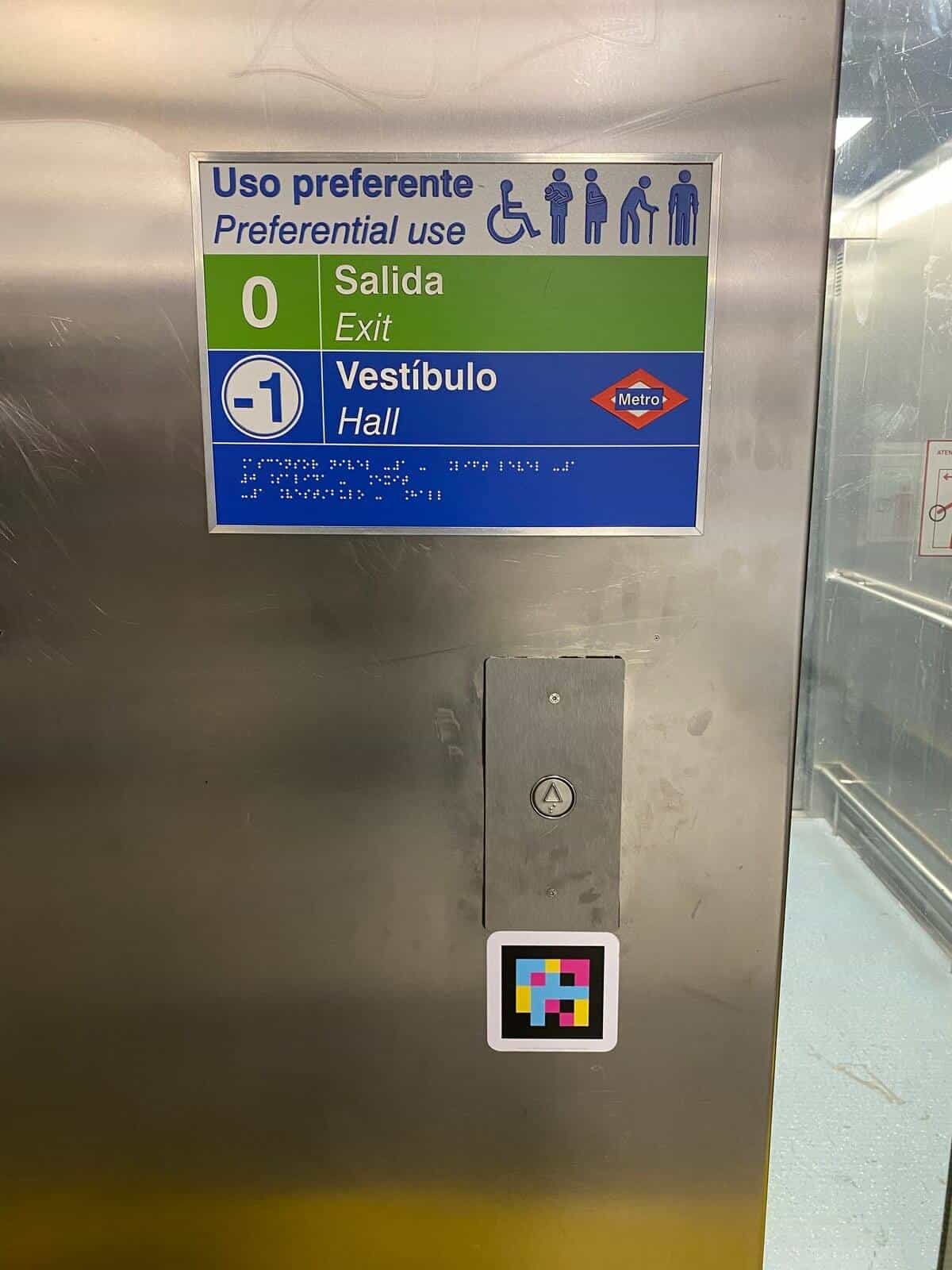 Inside a lift at Colombia station, Metro de Madrid: “Priority use” panel with pictograms of people with disabilities, levels “0 Exit” and “−1 Concourse” with the Metro's red diamond and embossed braille; underneath, the cabin button and a square NaviLens code at user height