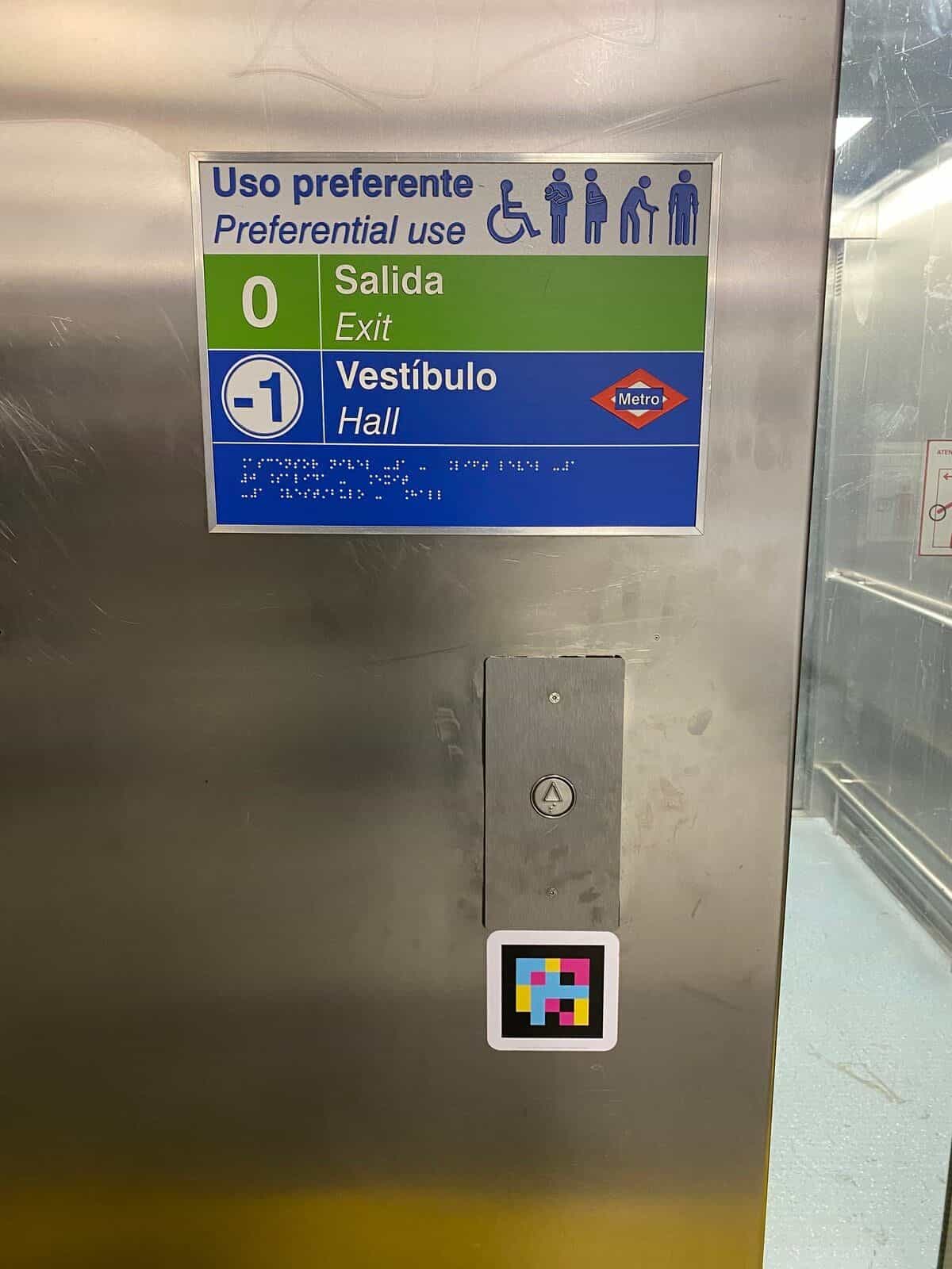 Inneres eines Aufzugs im Bahnhof Colombia von Metro de Madrid: Tafel „Vorrangnutzung“ mit Piktogrammen von Menschen mit Behinderung, Ebenen „0 Ausgang“ und „−1 Verteilerebene“ mit der roten Metro-Raute und Reliefbraille; darunter die Kabinentaste und ein quadratischer NaviLens-Code in Benutzerhöhe
