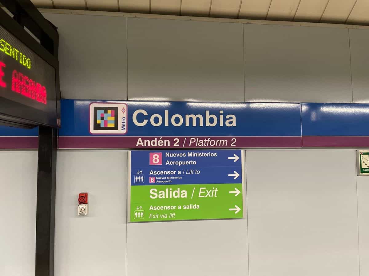 Detail des Bahnsteigschilds in Colombia: blaue Tafel „Colombia — Bahnsteig 2“ mit einem großen NaviLens-Code neben der roten Metro-Raute; darunter „8 Nuevos Ministerios — Flughafen“, „Aufzug zu“-Hinweise und das grüne Band „Ausgang — Aufzug zum Ausgang“