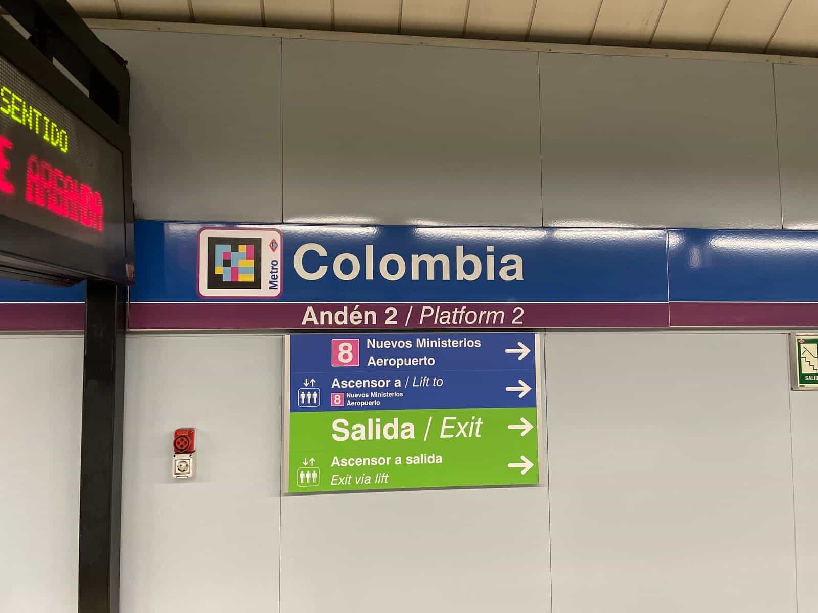 Colombiaのホーム標識のディテール。青いパネル「Colombia — 2番ホーム」に地下鉄の赤いダイヤモンドの隣に大きなNaviLensコード。下に「8 Nuevos Ministerios — 空港」、「エレベーターで」の表示、緑の帯「出口 — 出口へのエレベーター」