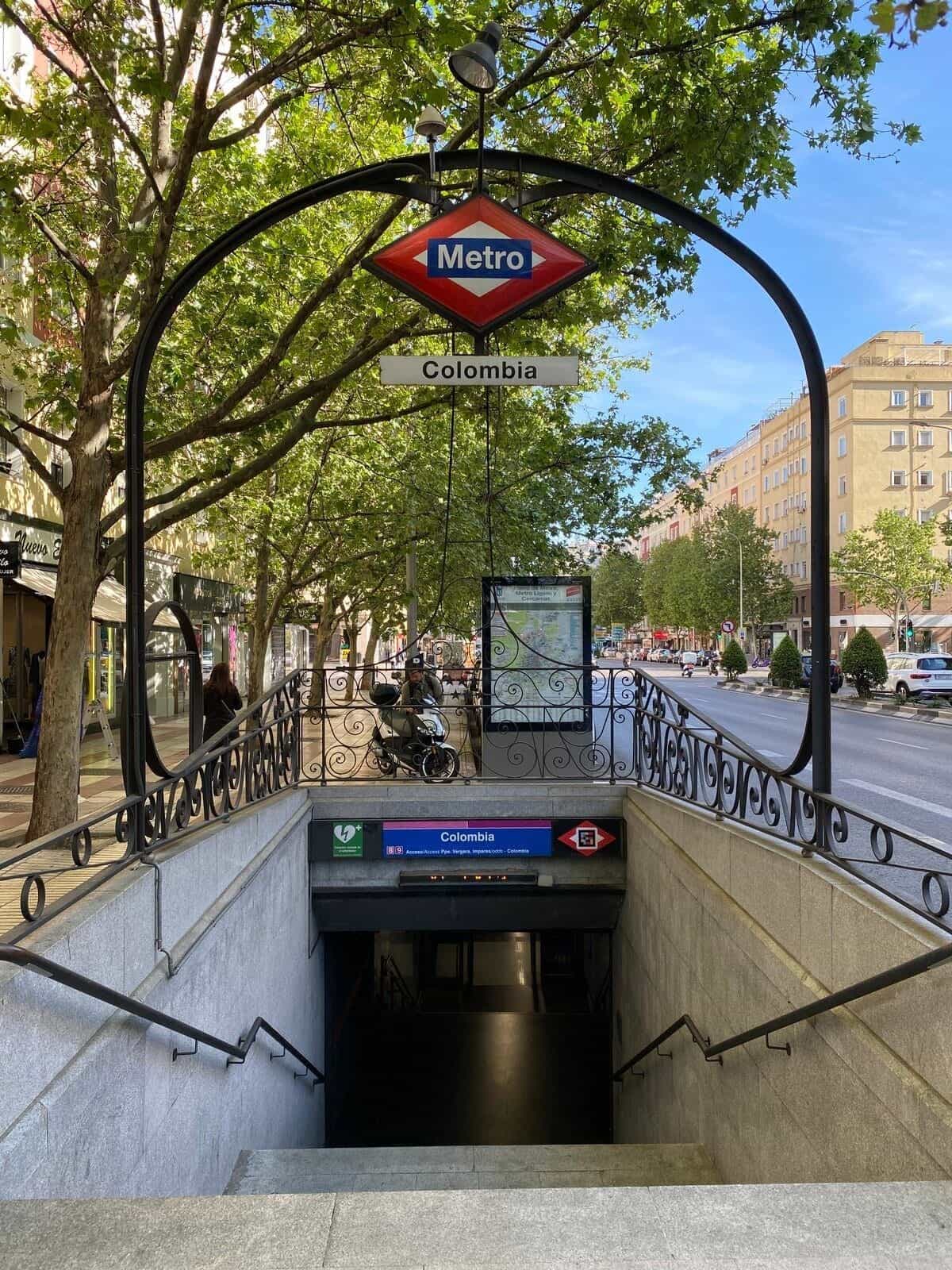 Außenzugang zum Bahnhof Colombia von Metro de Madrid: ein modernistischer schmiedeeiserner Bogen mit der roten Metro-Raute in der Mitte und dem hängenden „Colombia“-Schild, neben einer baumbestandenen Allee; im Hintergrund führen die Treppen zum Bahnhof hinunter, unter dem blauen Identifikationsschild mit dem NaviLens-Code