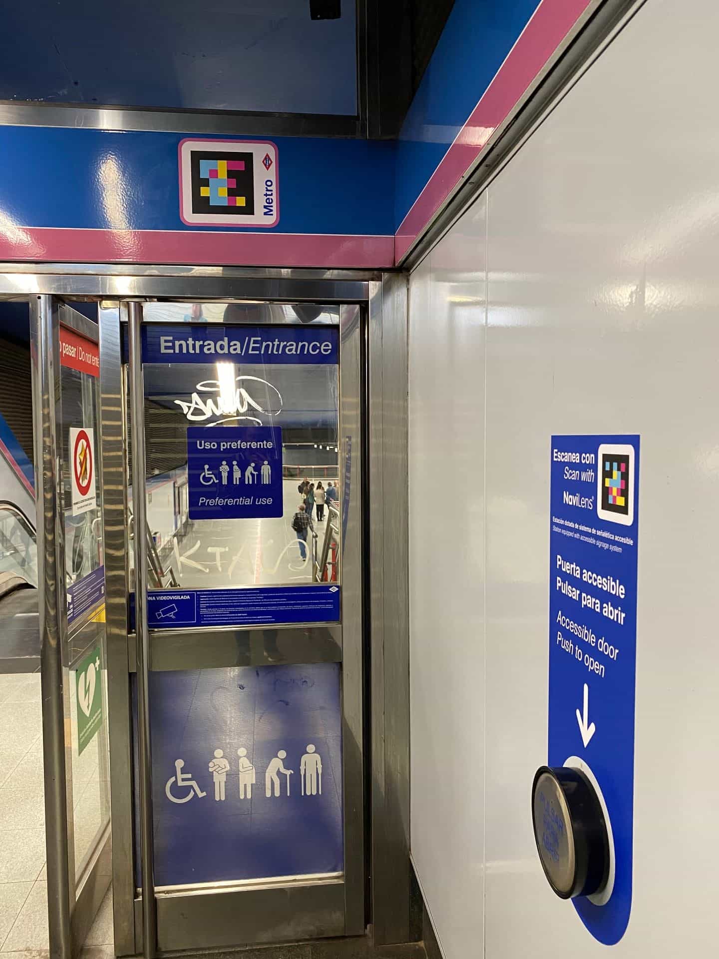 マドリード地下鉄Colombia駅のアクセシブルドア。上部、8号線の青とピンクのフレームに、地下鉄の赤いダイヤモンドの隣に額装されたNaviLensコード。通路の右側には青いパネル「NaviLensでスキャン — アクセシブル標識システムの駅 / アクセシブルドア — 押して開ける」と丸い青いボタンと下矢印