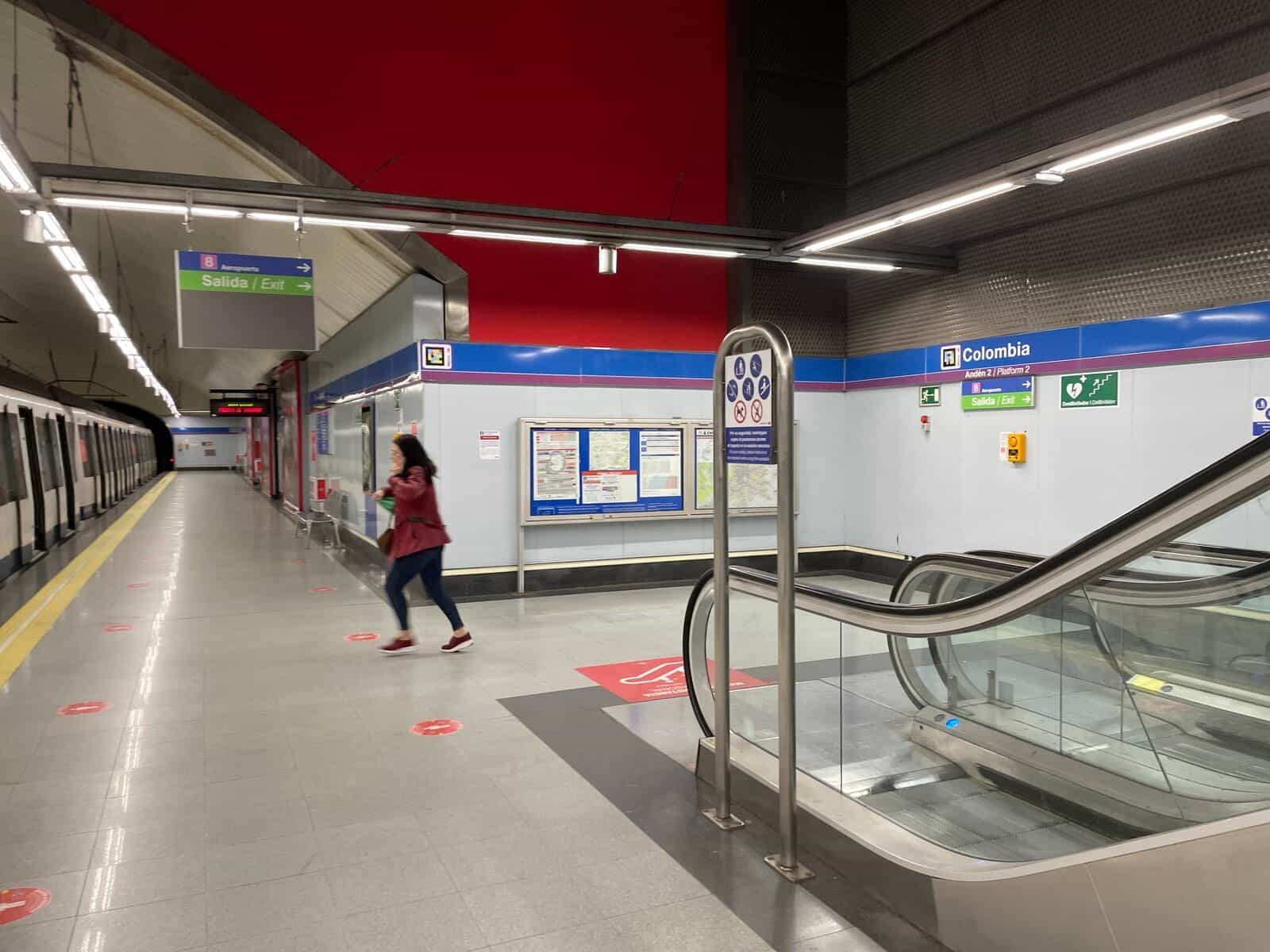 Colombia駅の中間階、右側にエスカレーター。天井から緑の「出口」と青の「8空港」標識が吊り下げられ、地下鉄路線図付きの情報パネルと、上端にNaviLensコード付きの「Colombia — 2番ホーム」標識