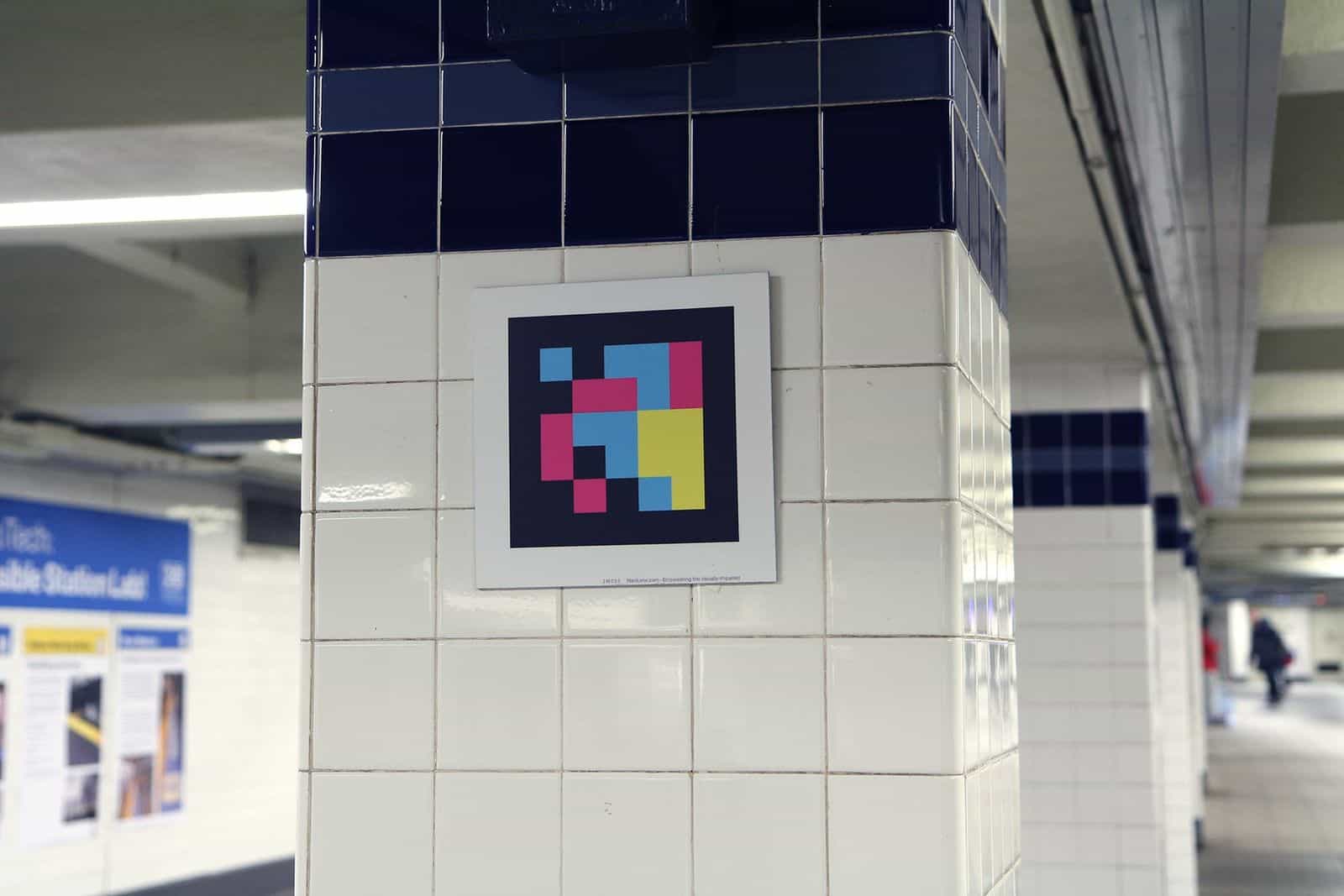 Bahnsteigsäule in Jay Street-MetroTech mit weißen Fliesen und einem marineblauen Fliesenband oben; mittig auf der Säulenfläche ein weiß gerahmter NaviLens-Code; im Hintergrund das blaue Panel «Accessible Station Lab»