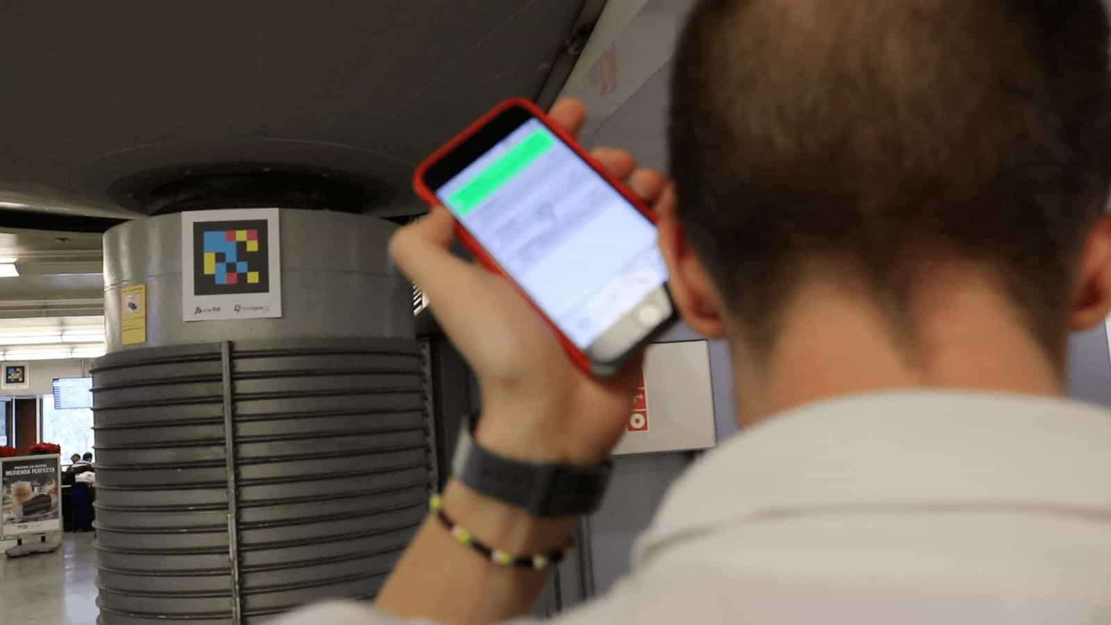 A round column in the Atocha concourse carries, at eye level, a square NaviLens code with the ‘Adif' and ‘NaviLens' logos below it; in the foreground, a traveler checks train information on the green screen of the NaviLens GO app on their phone.