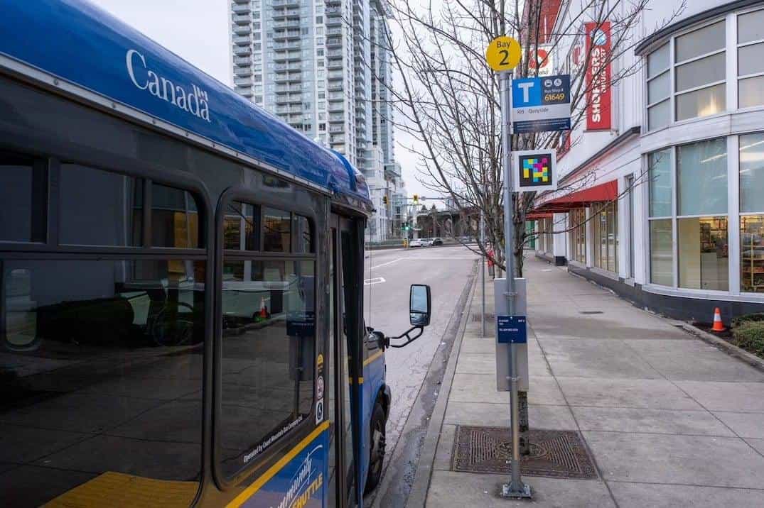 TransLink Community Shuttle (blue livery with the white “Canada” marking) at Bay 2 next to a stop pole with a yellow disc, “Bus Stop # 61649 — 103 Quayside” sign and a colourful NaviLens code framed below it