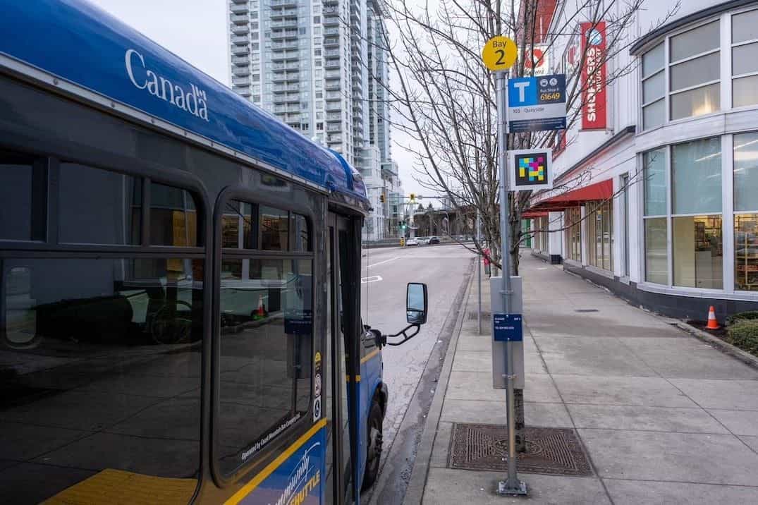 Community Shuttle de TransLink (carrocería azul con marca «Canada» en blanco) detenido en Bay 2 junto a un poste de parada con disco amarillo, cartel «Bus Stop # 61649 — 103 Quayside» y código NaviLens a colores enmarcado bajo el cartel