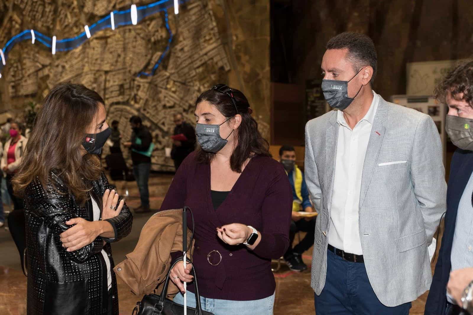 L’assessora María Navarro e un’utente della ONCE con mascherina del Gruppo Sociale ONCE conversano nella hall del municipio; sullo sfondo, il grande plastico-murale in rilievo di Saragozza con il corso dell’Ebro illuminato di blu