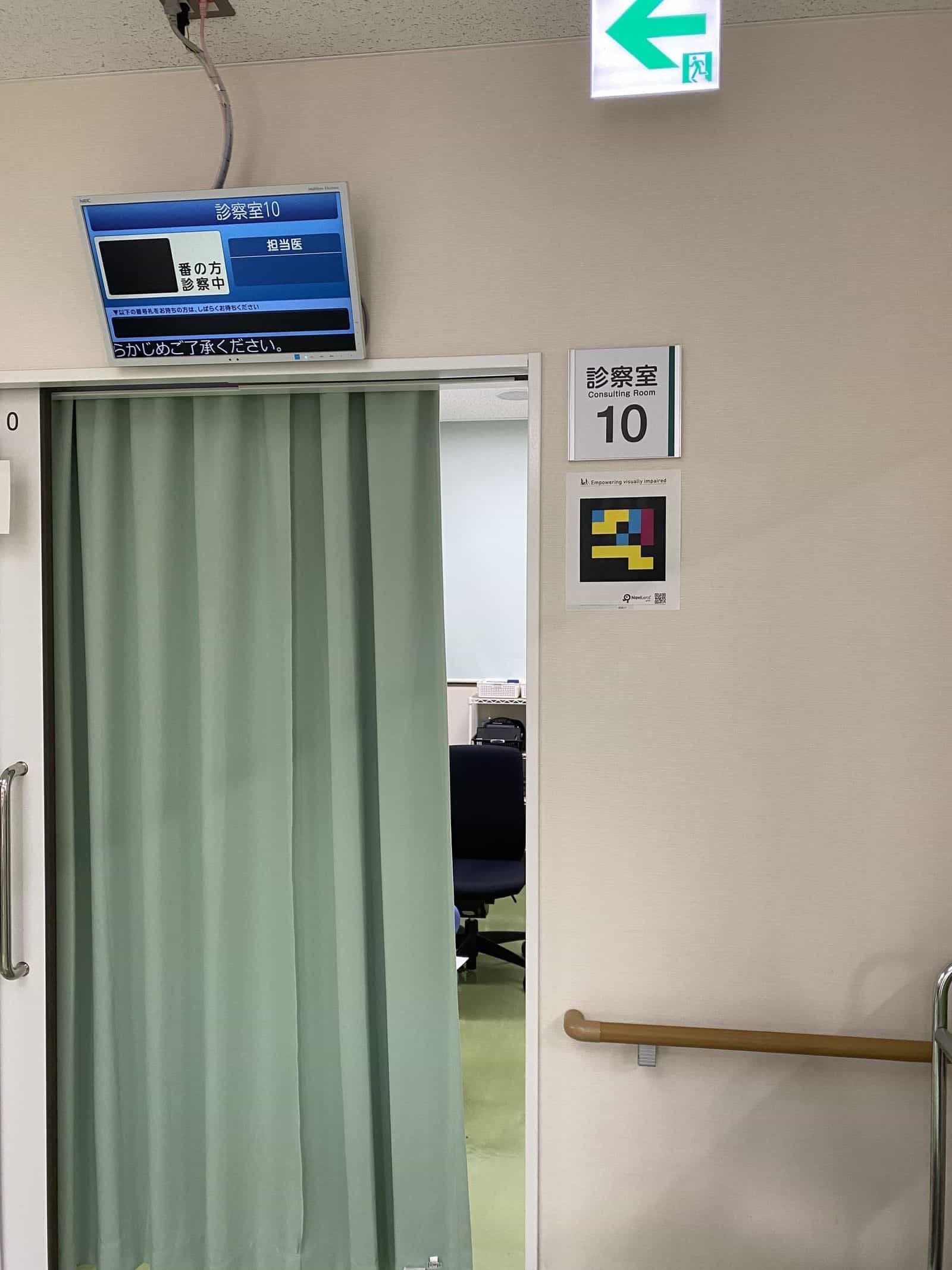 眼科 診察室10の扉：開口部にミントグリーンのカーテン、上部に青いNEC表示器（漢字で10）、右の壁に白い「診察室10」サインと「Empowering visually impaired」と書かれたラミネート版NaviLensコード、奥に医師の椅子と机が見える