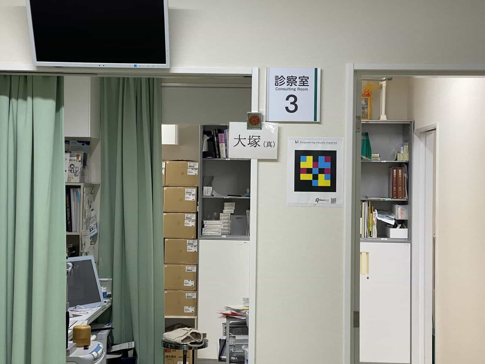 診察室3の扉：「診察室 / Consulting Room 3」パネルと「大塚（真）」と書かれた白い医師札、その右側にPublic Kitのラミネート版NaviLensコードがクリーム色の壁に透明テープで貼られている