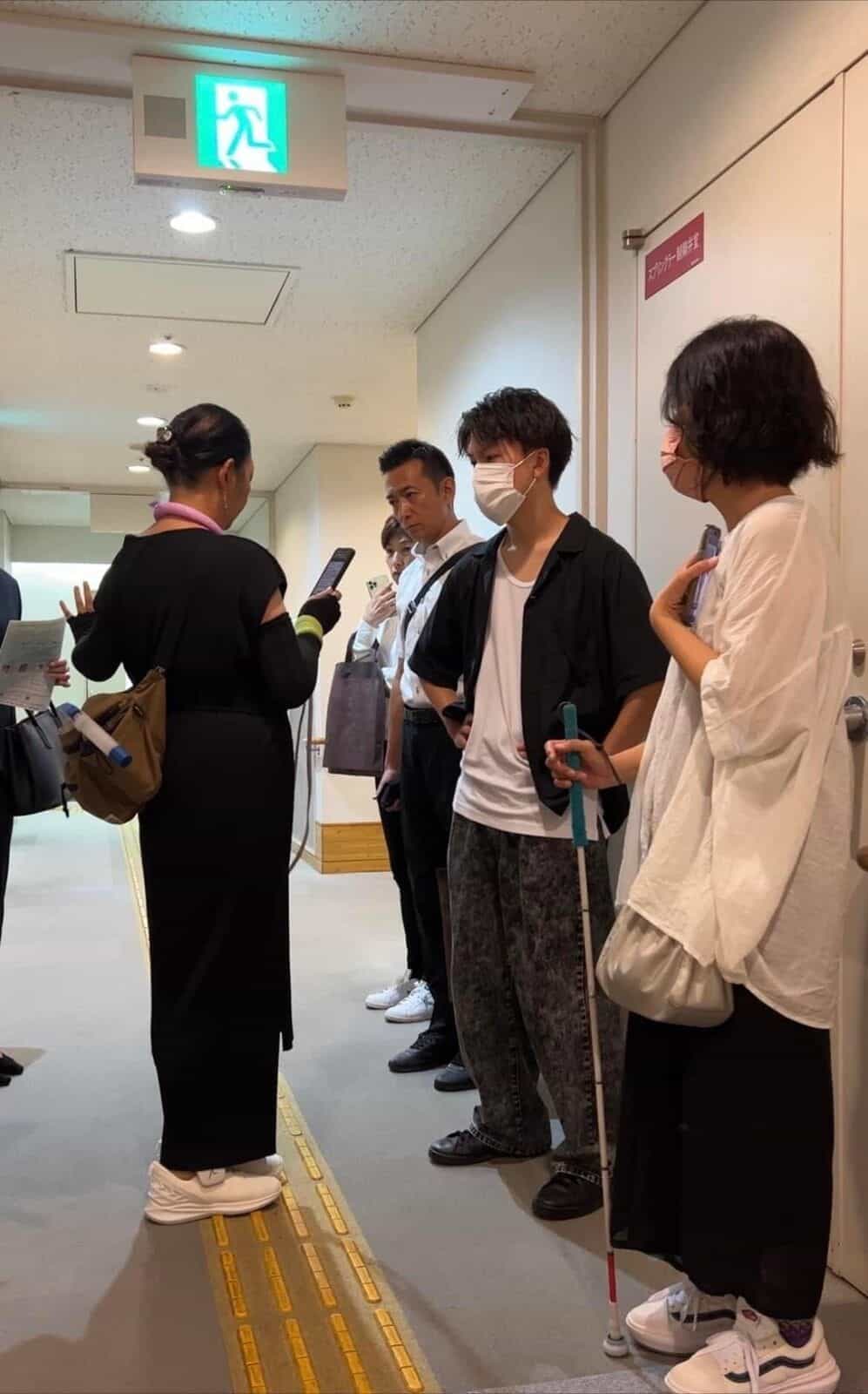 廊下で NaviLens 案内板の横でメジンスの説明を聞くまゆだまネットフェスタの参加者グループ