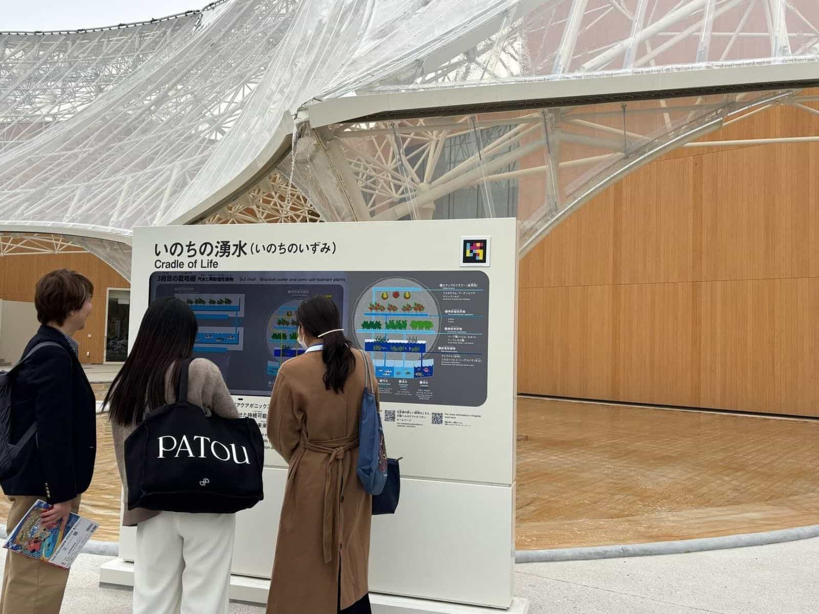 大阪ヘルスケアパビリオンの「いのちの湧水・Cradle of Life」パネル、汽水でのアクアポニクス栽培の図解と右上に設置されたNaviLensコード。3人の来場者がパネルを読んでいる