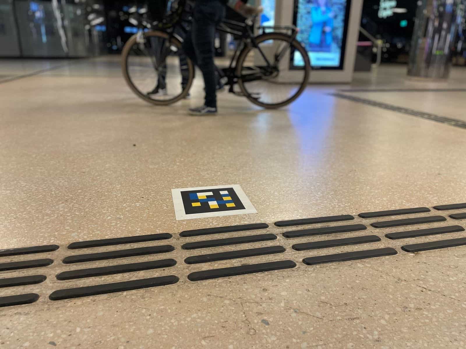 NaviLens-Code im Boden der Halle von Amsterdam Centraal neben schwarzen taktilen Leitlinien, im Hintergrund eine Person, die ein Fahrrad schiebt