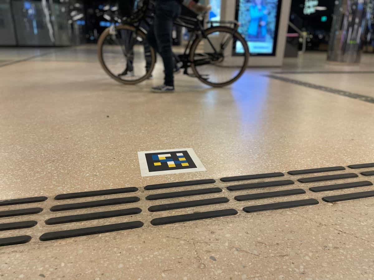 NaviLens-Code im Boden der Halle von Amsterdam Centraal neben schwarzen taktilen Leitlinien, im Hintergrund eine Person, die ein Fahrrad schiebt