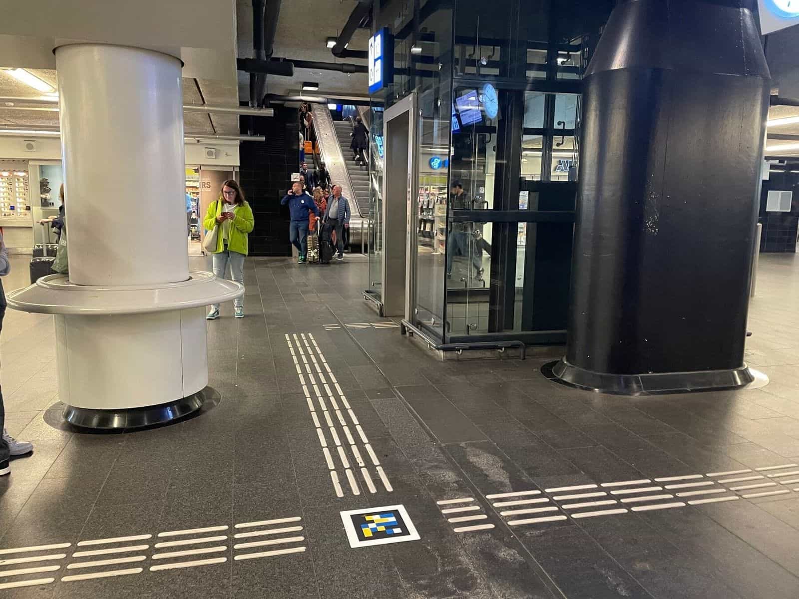 Innenkorridor von Amsterdam Centraal mit taktilen Leitlinien und einem farbigen NaviLens-Code im Boden, neben dem Aufzug