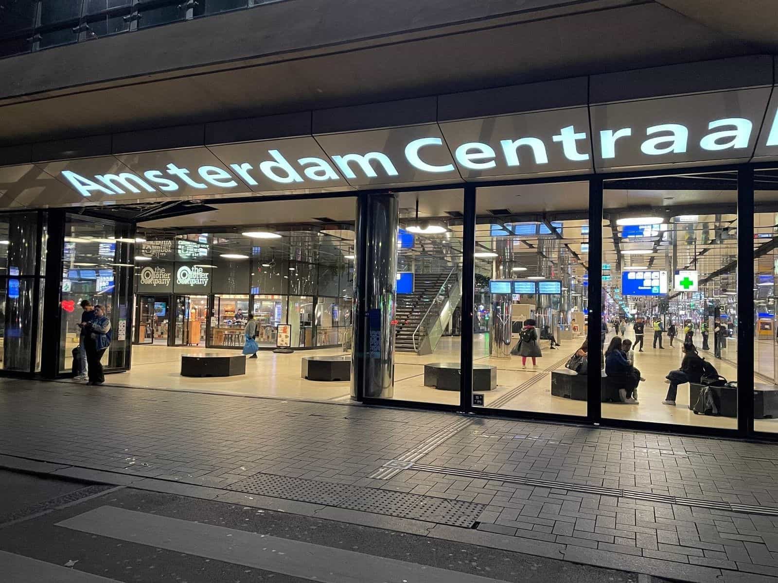 Beleuchtete Fassade von Amsterdam Centraal bei Nacht mit dem Schriftzug 'Amsterdam Centraal' in großen weißen Buchstaben und Reisenden, die in den Bahnhof gehen