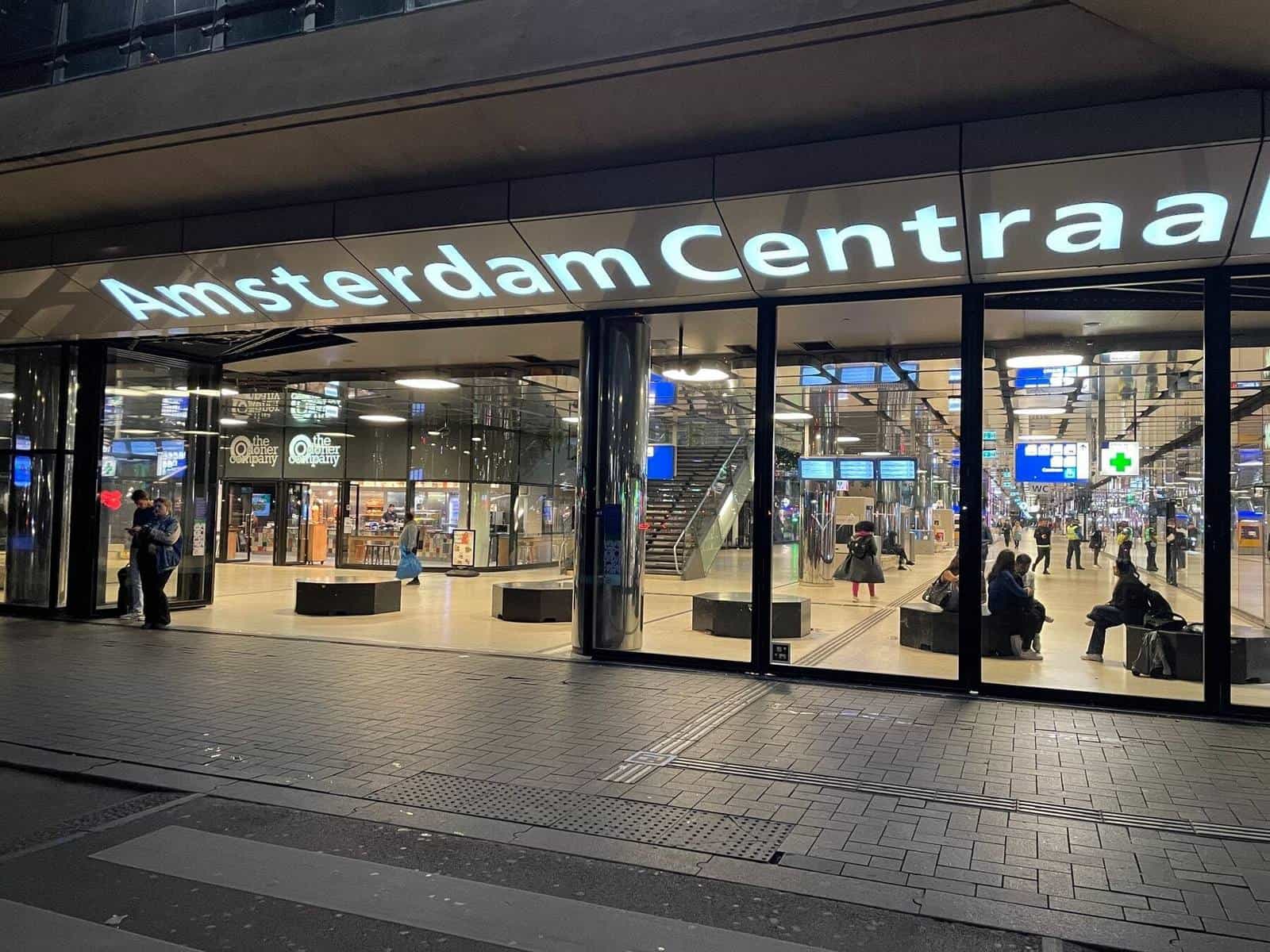 Illuminated façade of Amsterdam Centraal at night with the 'Amsterdam Centraal' sign in large white letters and travellers entering the station