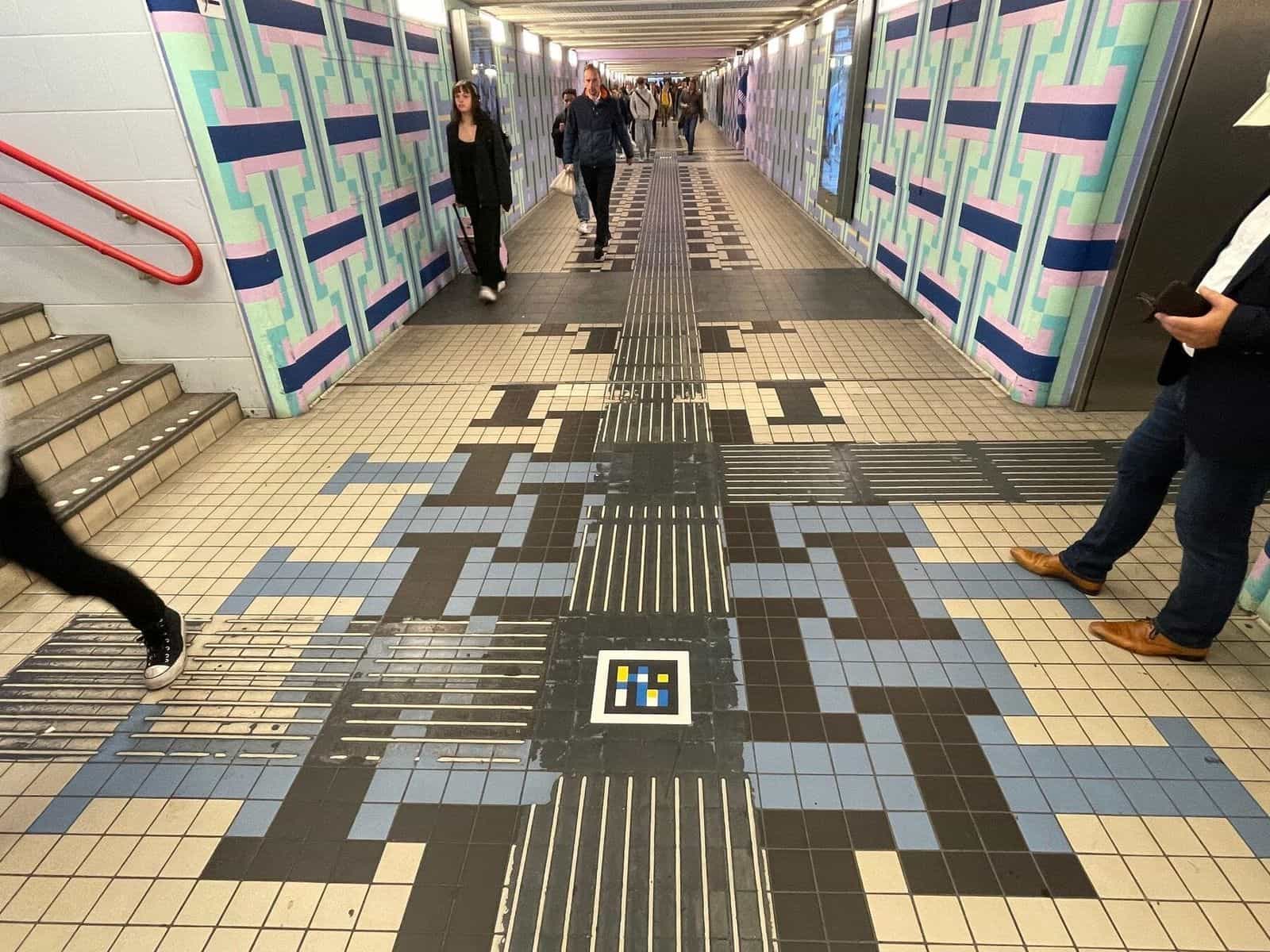 Durchgangstunnel in Amsterdam Centraal mit geometrischem Mosaikboden, mittigen Leitlinien und einem NaviLens-Code an einer Kreuzung
