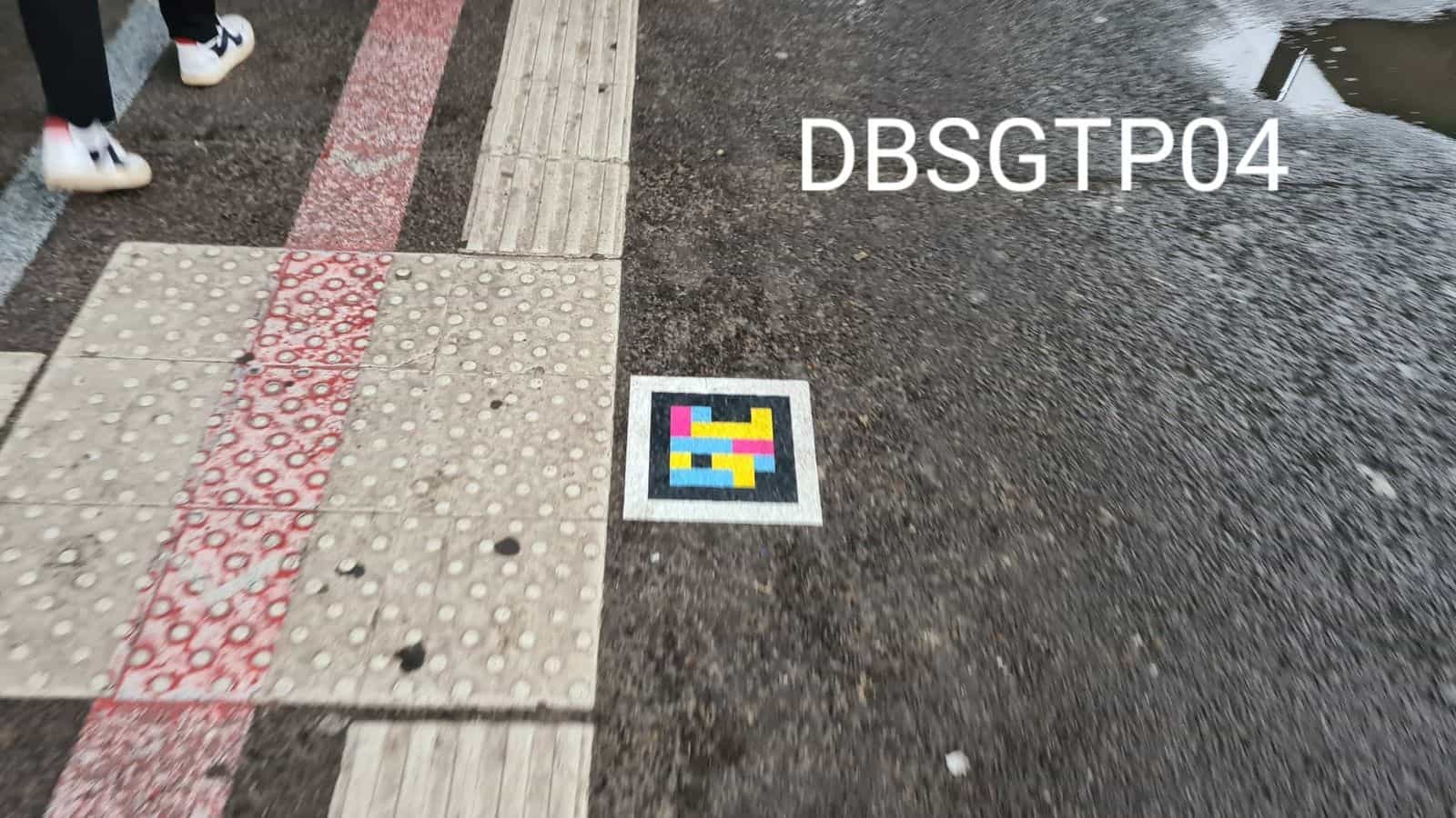 Asphalte mouillé près du Hbf avec dalle podotactile, bande rouge de piste cyclable et code NaviLens carré collé sur l’asphalte réfléchissant (DBSGTP04)
