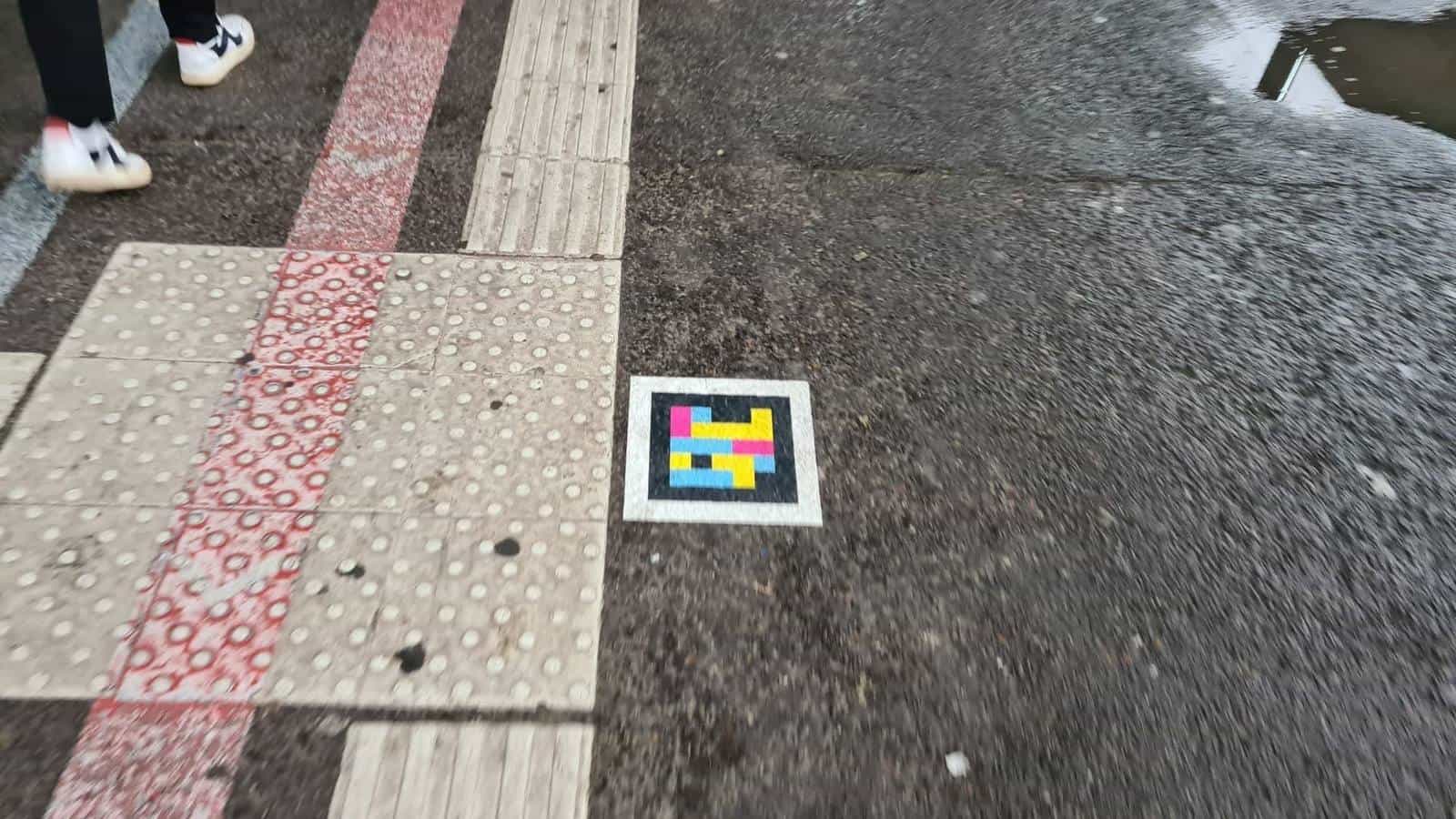 Wet asphalt next to the Hbf with a tactile slab, a red bike-lane strip and a NaviLens square code stuck on the reflective asphalt