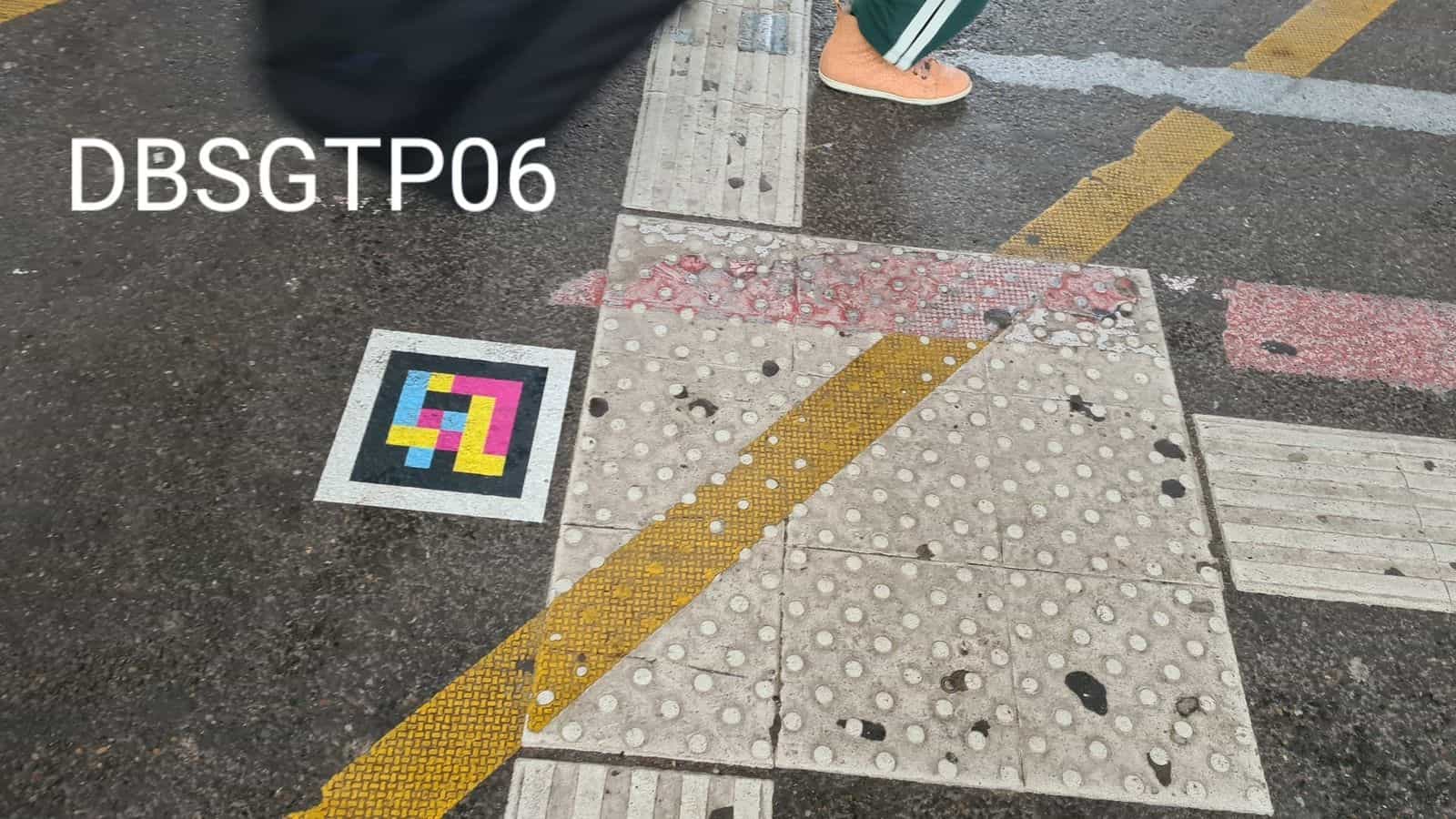 Passage piéton au Hbf avec dalles podotactiles et bandes jaunes/rouges ; un passant traverse à côté d’un code NaviLens collé sur l’asphalte (DBSGTP06)