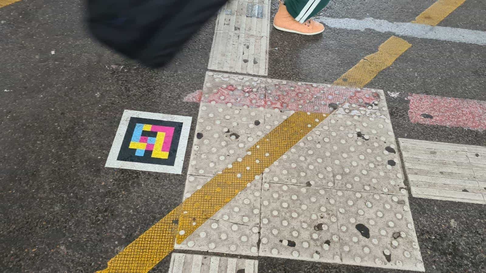 Pedestrian crossing at the Hbf with tactile slabs and yellow/red strips; a pedestrian walks past a NaviLens code stuck to the asphalt