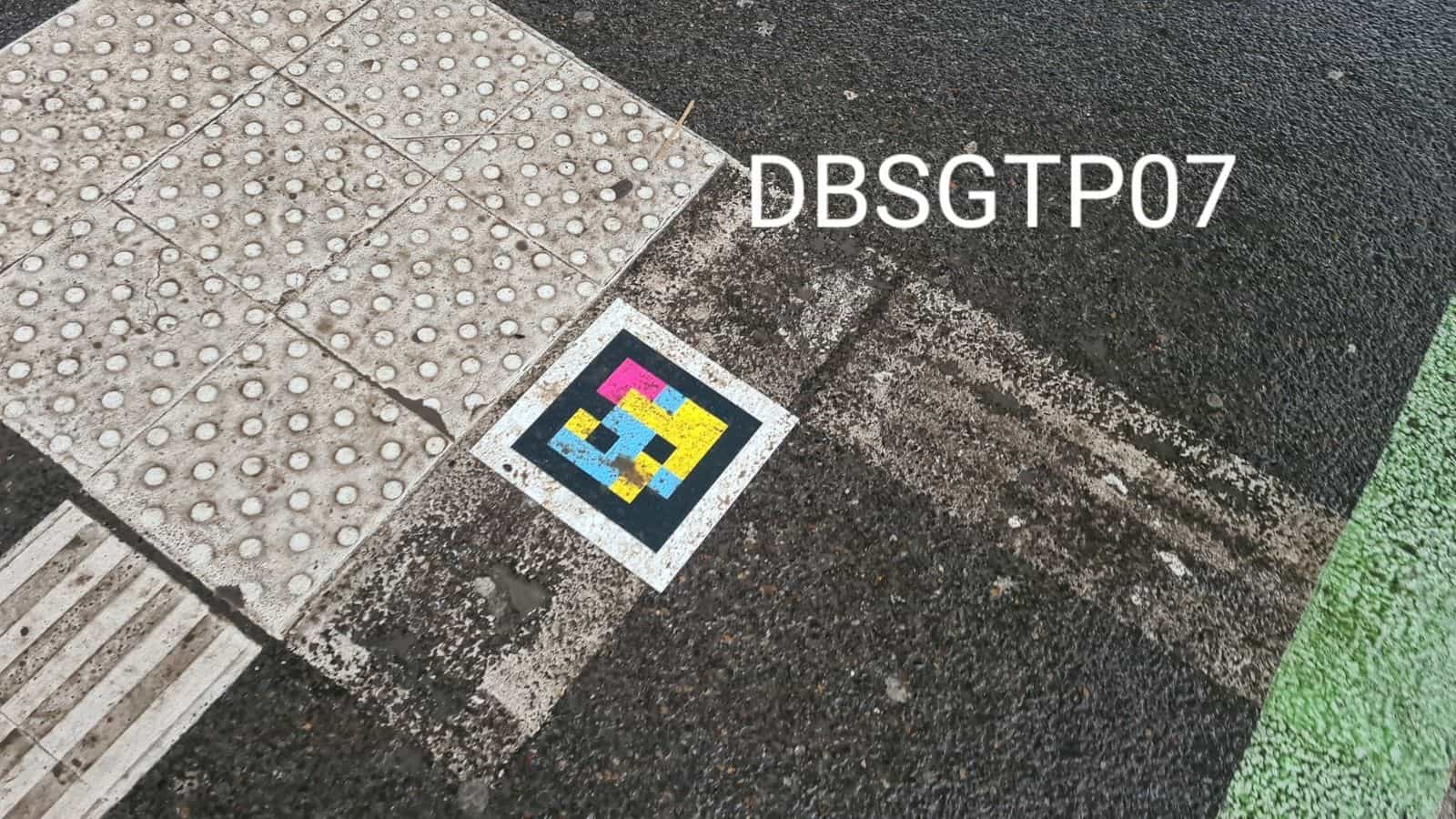 Coin d’un carrefour au Hbf avec dalle podotactile, asphalte mouillé et bande de gazon synthétique vert à côté d’un code NaviLens (DBSGTP07)