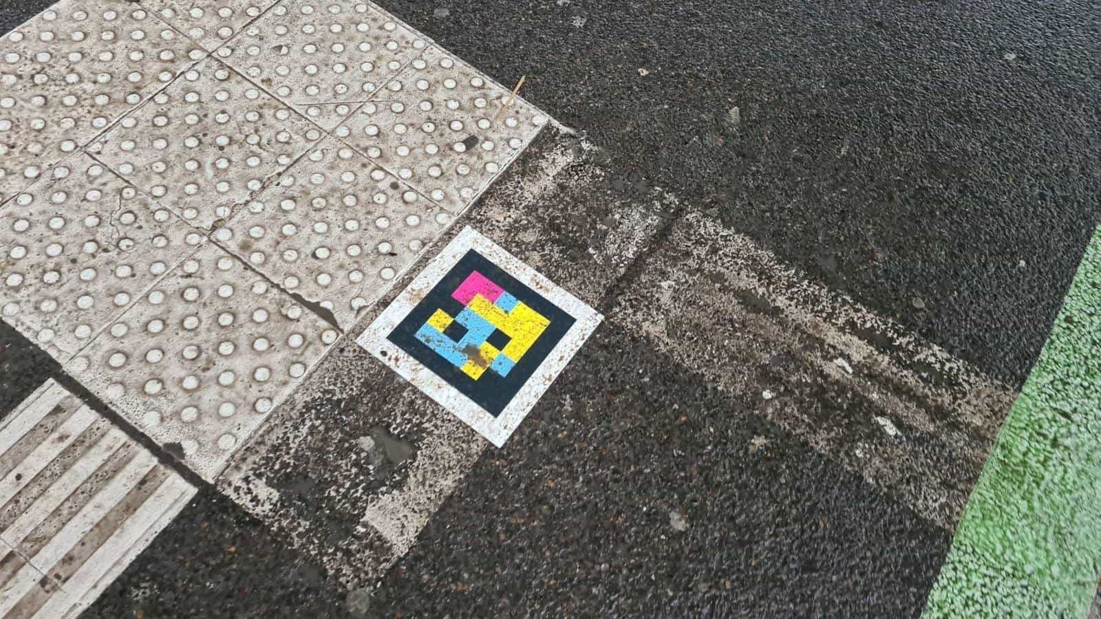 Corner of a crossing at the Hbf with tactile slab, wet asphalt and a strip of green artificial grass next to a NaviLens code