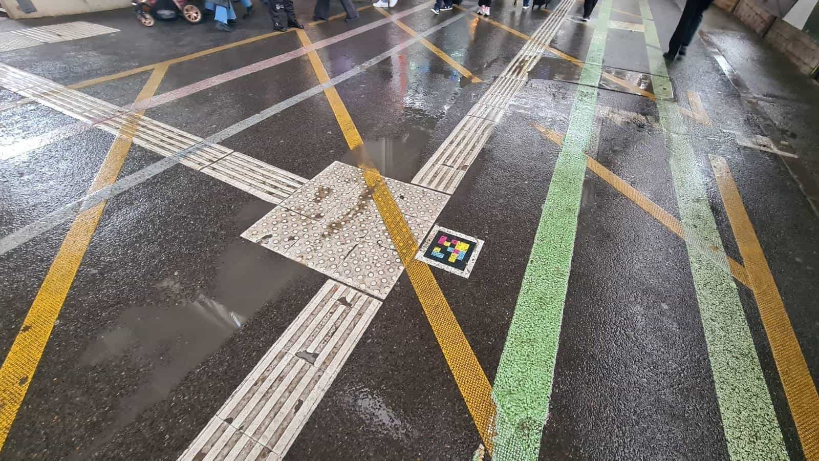 Top-down view of the floor of the Klett-Passage in Stuttgart in the rain: crossing of white tactile guide lines and yellow and green strips meeting at a dotted attention tile with a square NaviLens code stuck on top