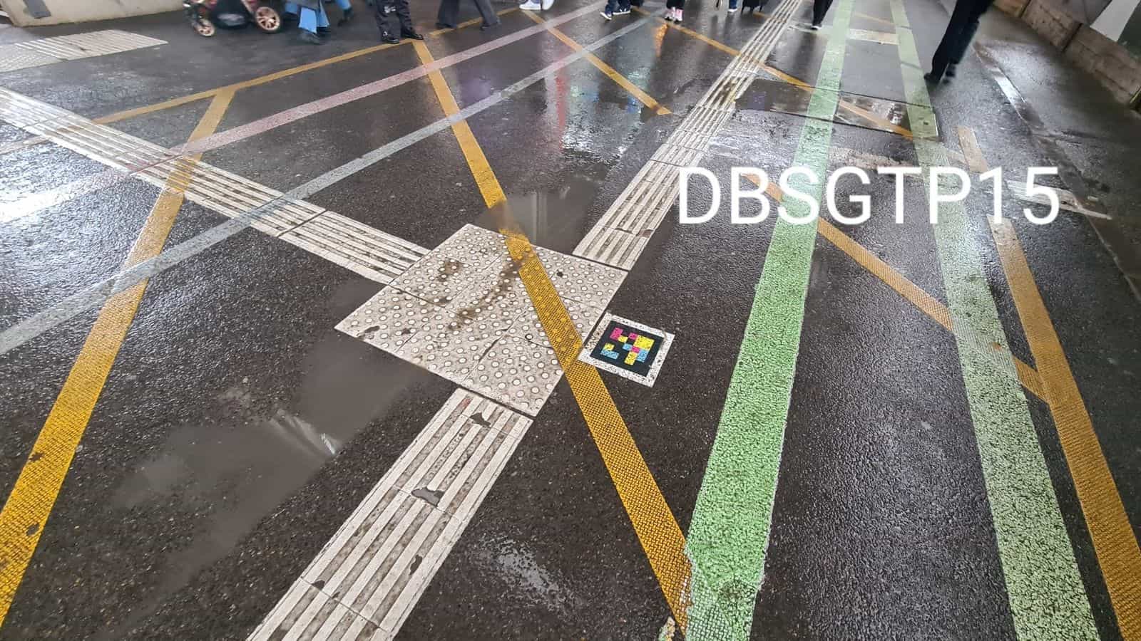 Vue plongeante du sol de la Klett-Passage de Stuttgart sous la pluie : croisement de bandes de guidage podotactiles blanches et de bandes jaunes et vertes convergeant vers une dalle à pastilles sur laquelle est collé un code NaviLens carré