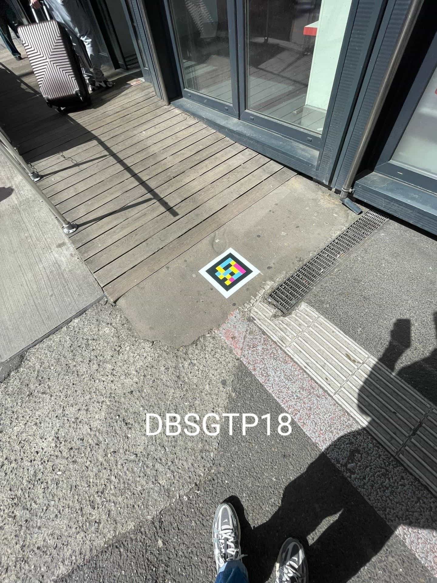 Vue large du même accès DBSGTP18 : une personne tire une valise rose sur le parquet pendant que le code NaviLens reste visible au sol