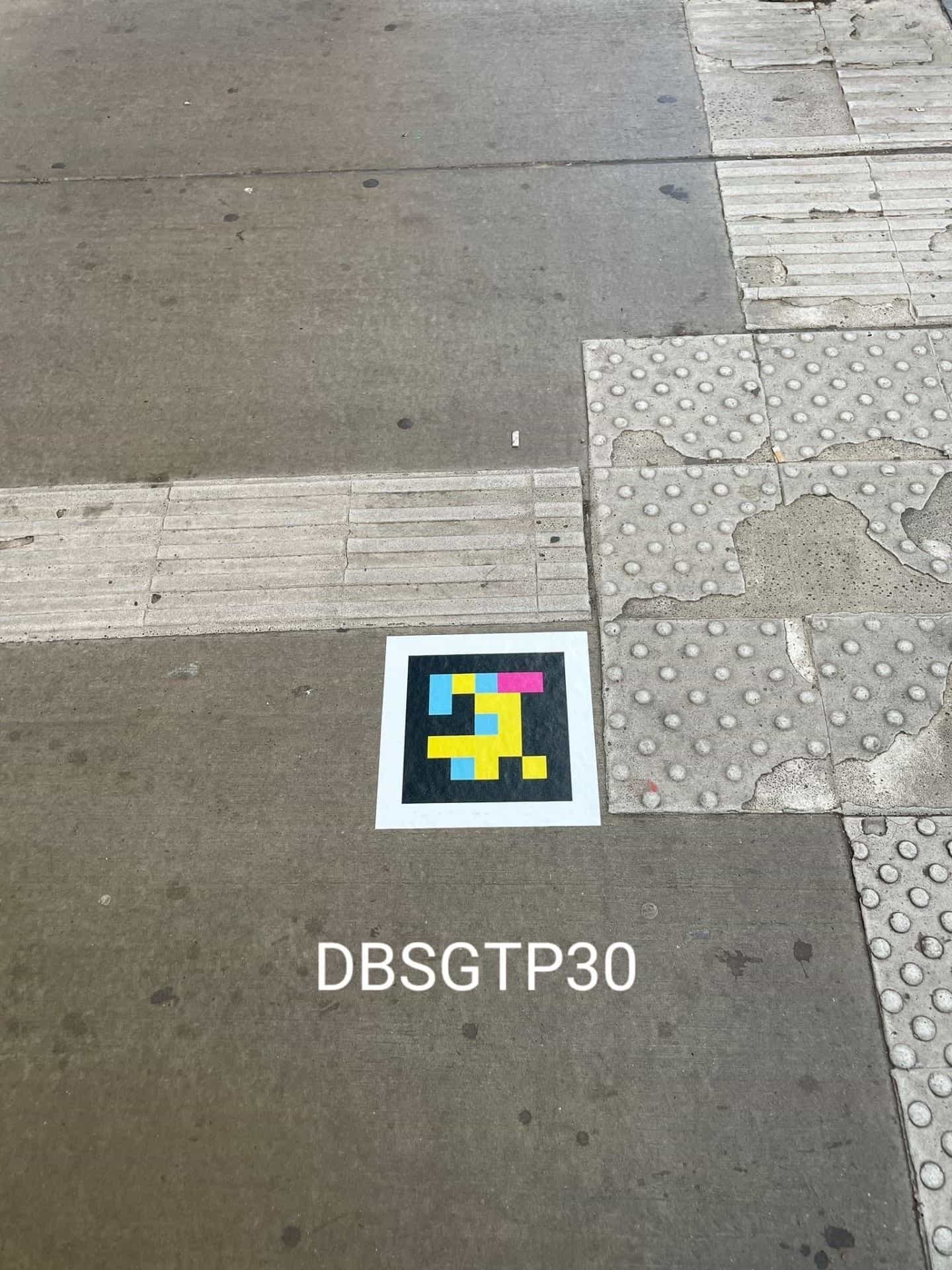 Trottoir en béton devant le Hbf avec dalles podotactiles et bandes de canalisation ; au centre, un code NaviLens carré collé au sol (DBSGTP30)