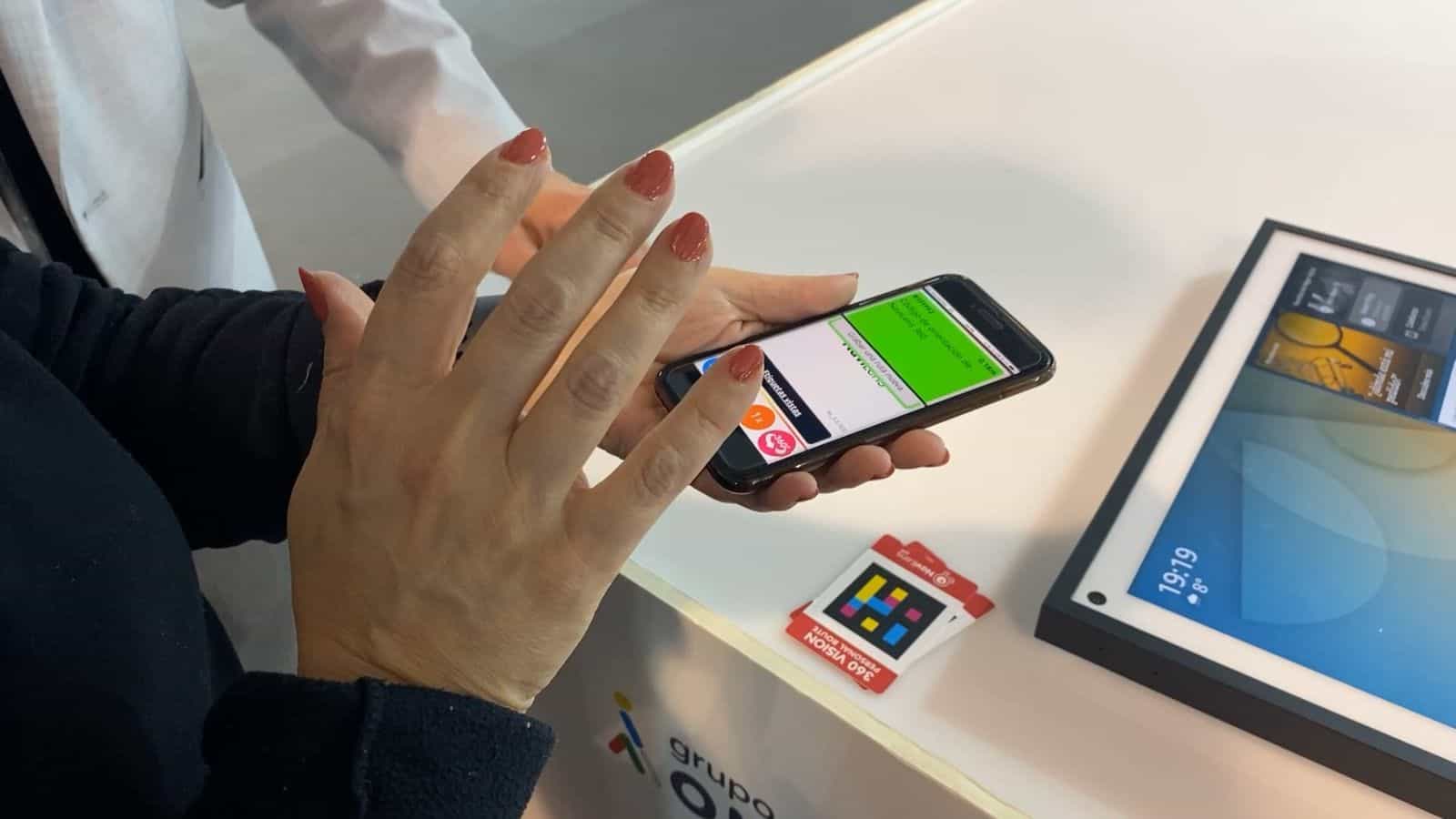 Demo allo stand del Grupo ACCEDO durante Tifloinnova 2022: le mani di una donna con le unghie smaltate di marrone tengono un iPhone in orizzontale il cui schermo mostra «Codice di orientamento di NaviLens 360 — 0,16 m, condividi nuovo percorso» in una fascia verde; accanto al telefono, una card pieghevole «360 VISION · Personal Route» con il codice NaviLens visibile
