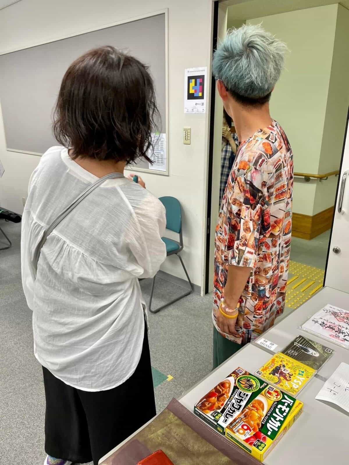 NaviLens ワークショップのデモ:群馬県社会福祉総合センターの廊下で NaviLens 案内板付きのドアの前に立つ2名