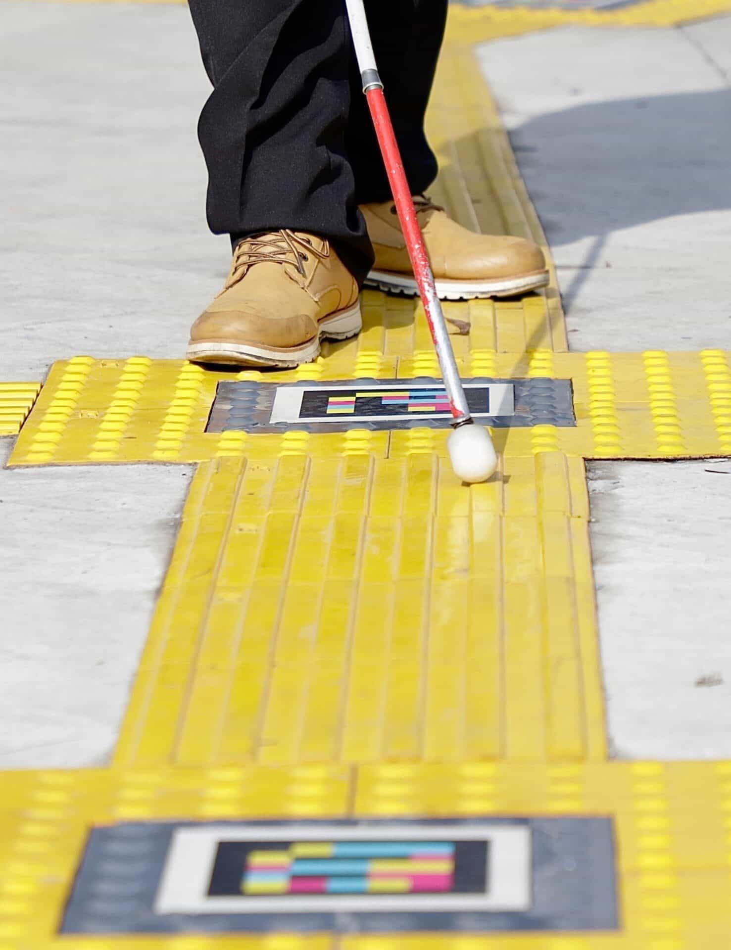 Close-up of feet in boots and a red-and-white cane crossing a node of tactile tiles with two NaviLens codes marking the direction of travel in Zapopan