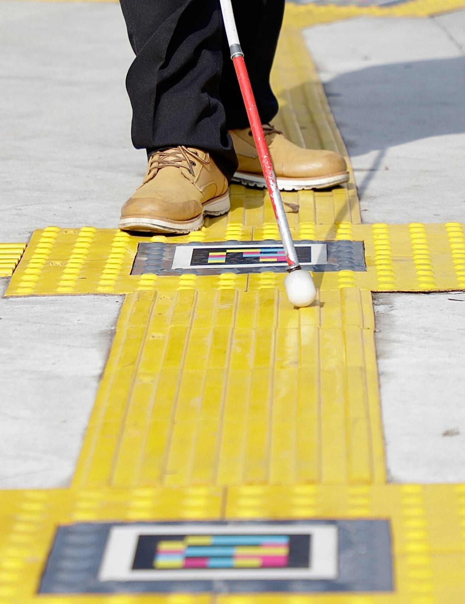 Close-up of feet in boots and a red-and-white cane crossing a node of tactile tiles with two NaviLens codes marking the direction of travel in Zapopan