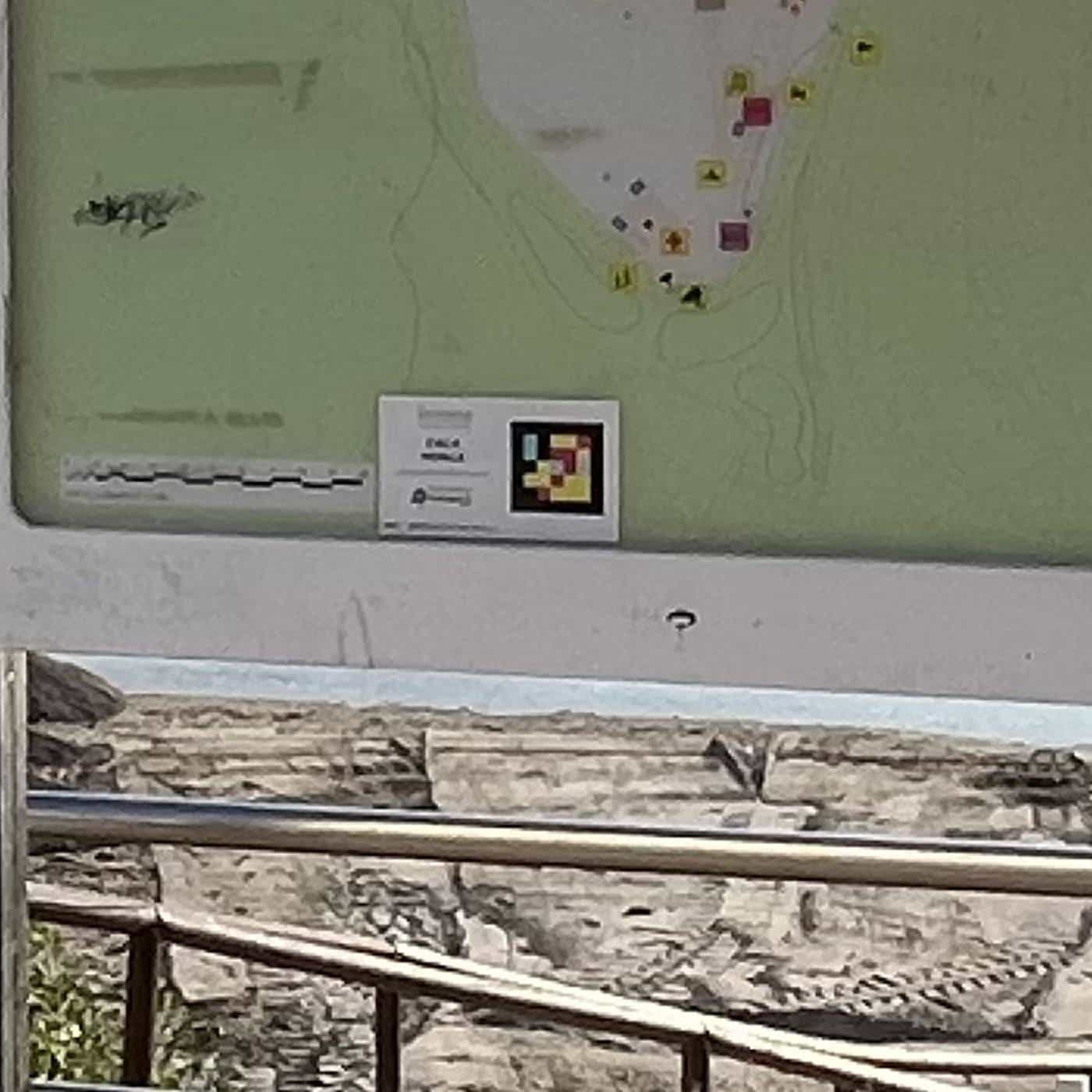 Close-up of the Cala Mosca location-map panel: over the map, a white plate with the beach name and the NaviLens code in yellow, red and turquoise on black, with the app label underneath; behind it, the rocky shore and the sea
