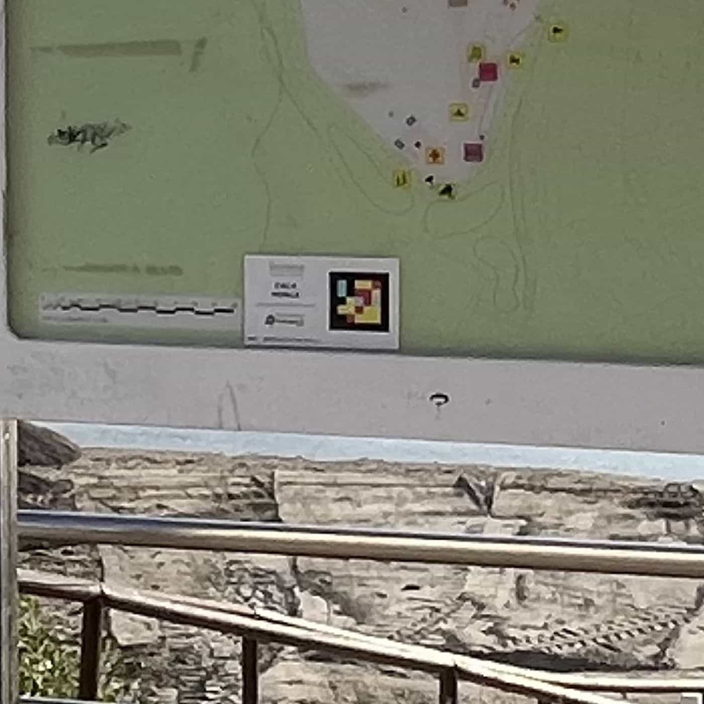 Detail der Lageplantafel von Cala Mosca: über der Karte ein weißes Schild mit dem Strandnamen und dem NaviLens-Code in Gelb, Rot und Türkis auf Schwarz, darunter der Hinweis auf die App; dahinter die Uferbefestigung und das Meer