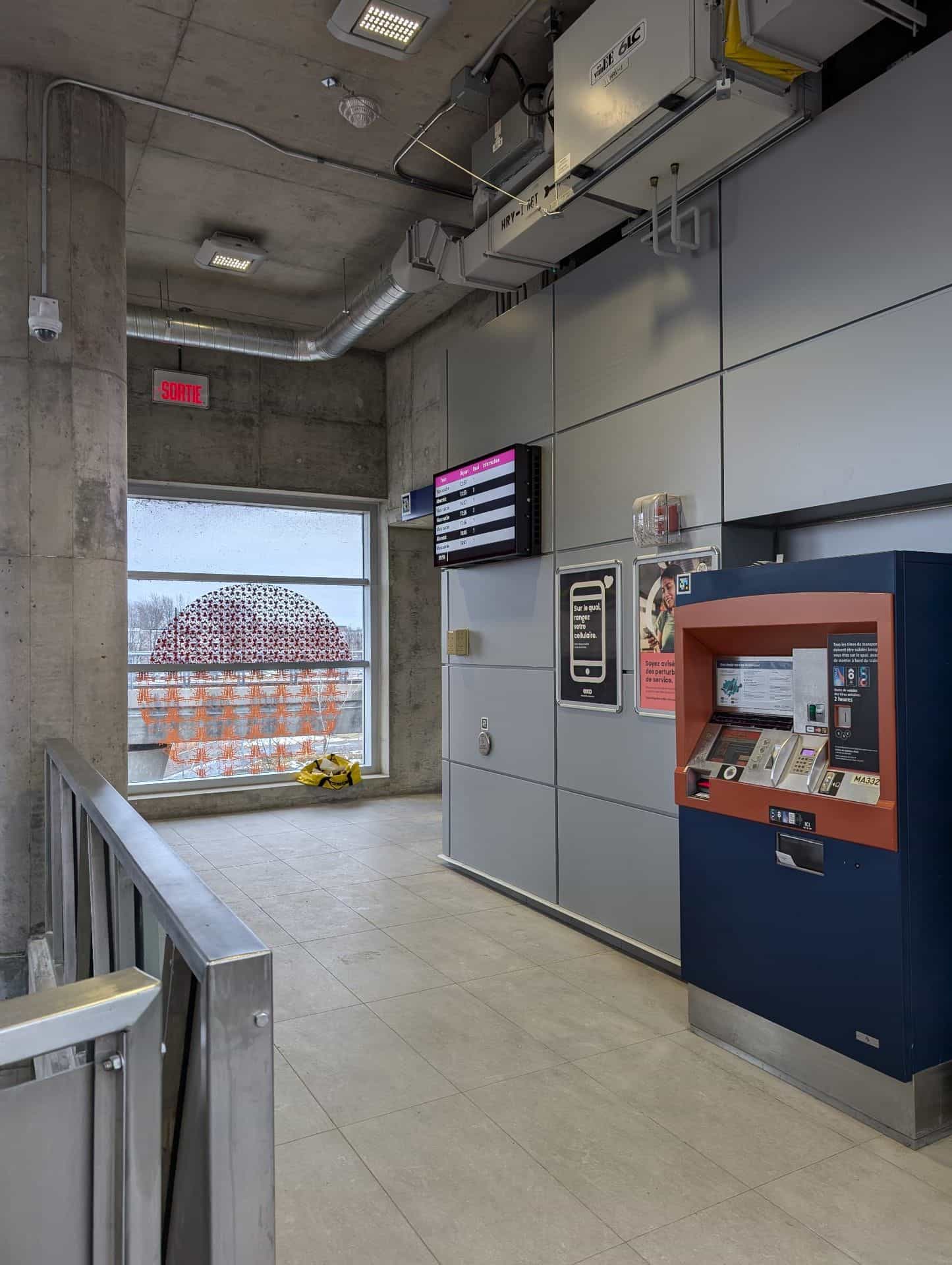 Raised concourse at an exo station with a navy-and-orange ticket machine and an LED screen showing upcoming departures to Mascouche and Ahuntsic; among the information posters, a NaviLens code in cyan, magenta and yellow