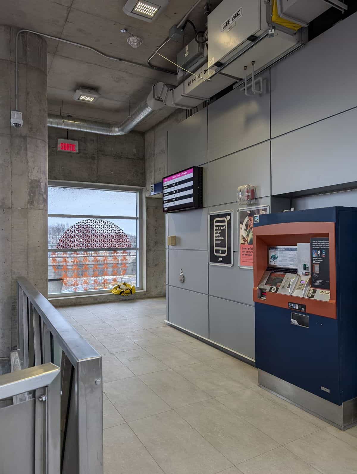 Raised concourse at an exo station with a navy-and-orange ticket machine and an LED screen showing upcoming departures to Mascouche and Ahuntsic; among the information posters, a NaviLens code in cyan, magenta and yellow