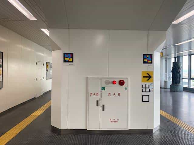 Corridor at Kobe Airport Station with two framed NaviLens tags on the white wall, next to a red and white fire-extinguisher cabinet and a yellow “Exit →” sign. On the floor, a continuous strip of yellow tactile paving