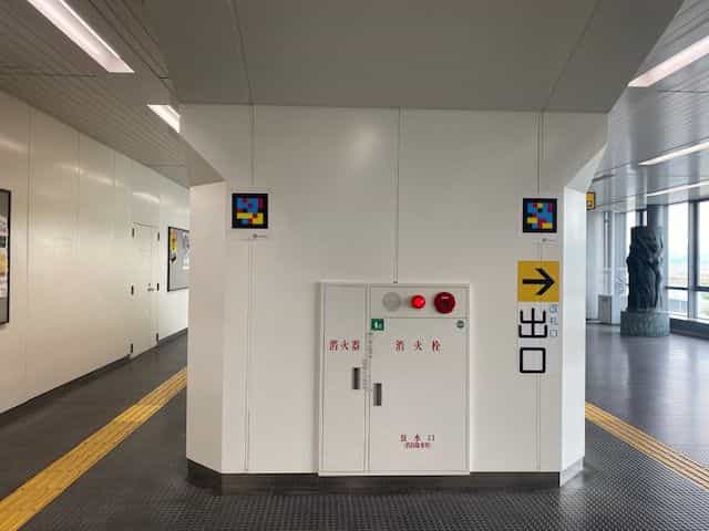 神戸空港駅の通路。白い壁に枠付きNaviLensタグが2つ、赤白の消火器収納庫と黄色の「出口 →」サインの横に並ぶ。床には連続した黄色の点字ブロック